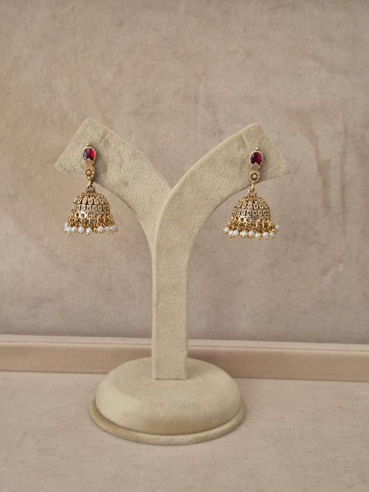 air of gold-toned Madhumita Jhumka earrings hanging on a beige velvet display stand, featuring teardrop-shaped studs with bright red gemstones and detailed bell-shaped drops fringed with small white pearls by naitri.in