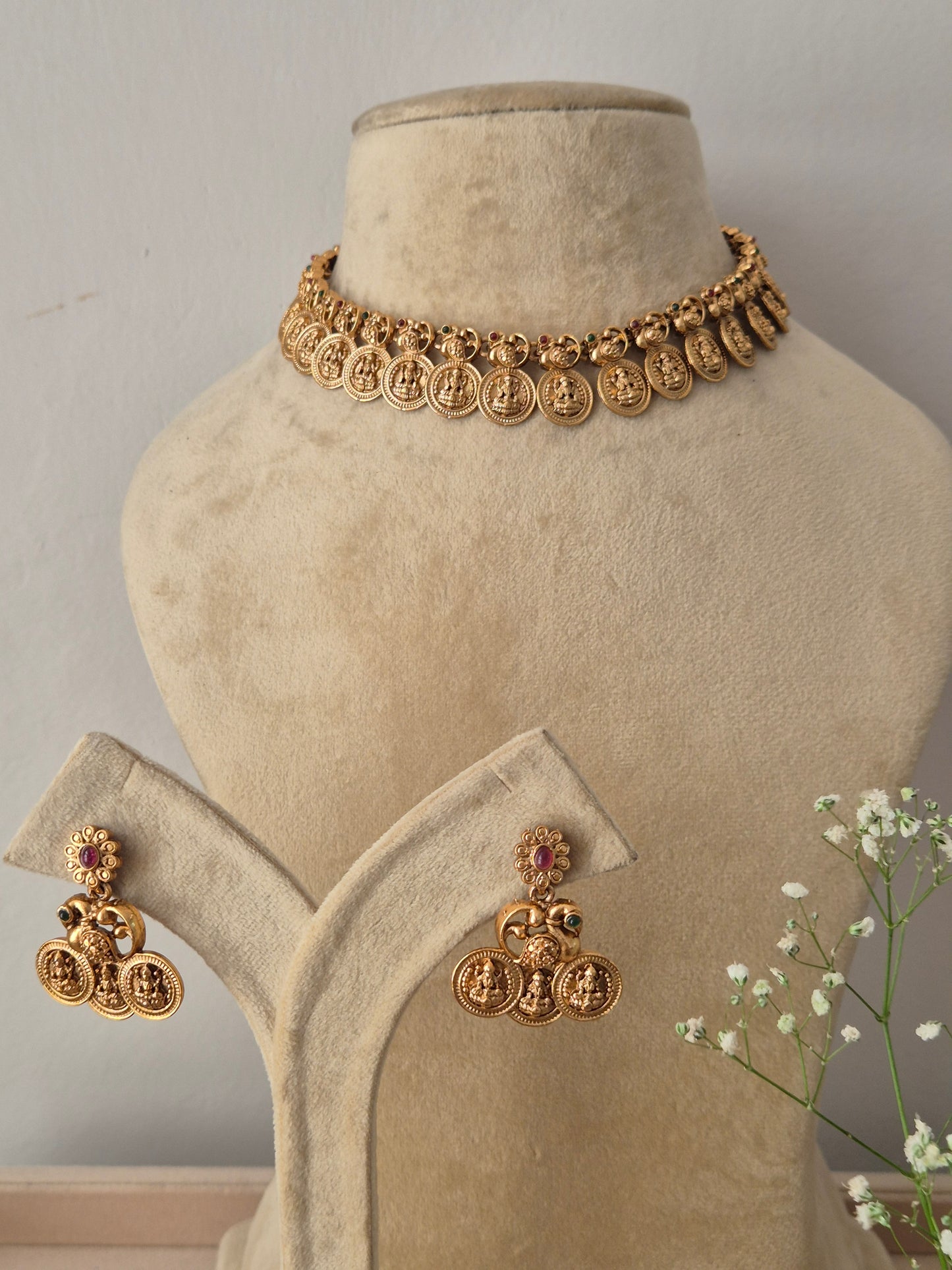 A gold-plated choker necklace featuring a repeating pattern of traditional coin-shaped motifs, with matching hanging jhumka earrings featuring pink accents by naitri.in