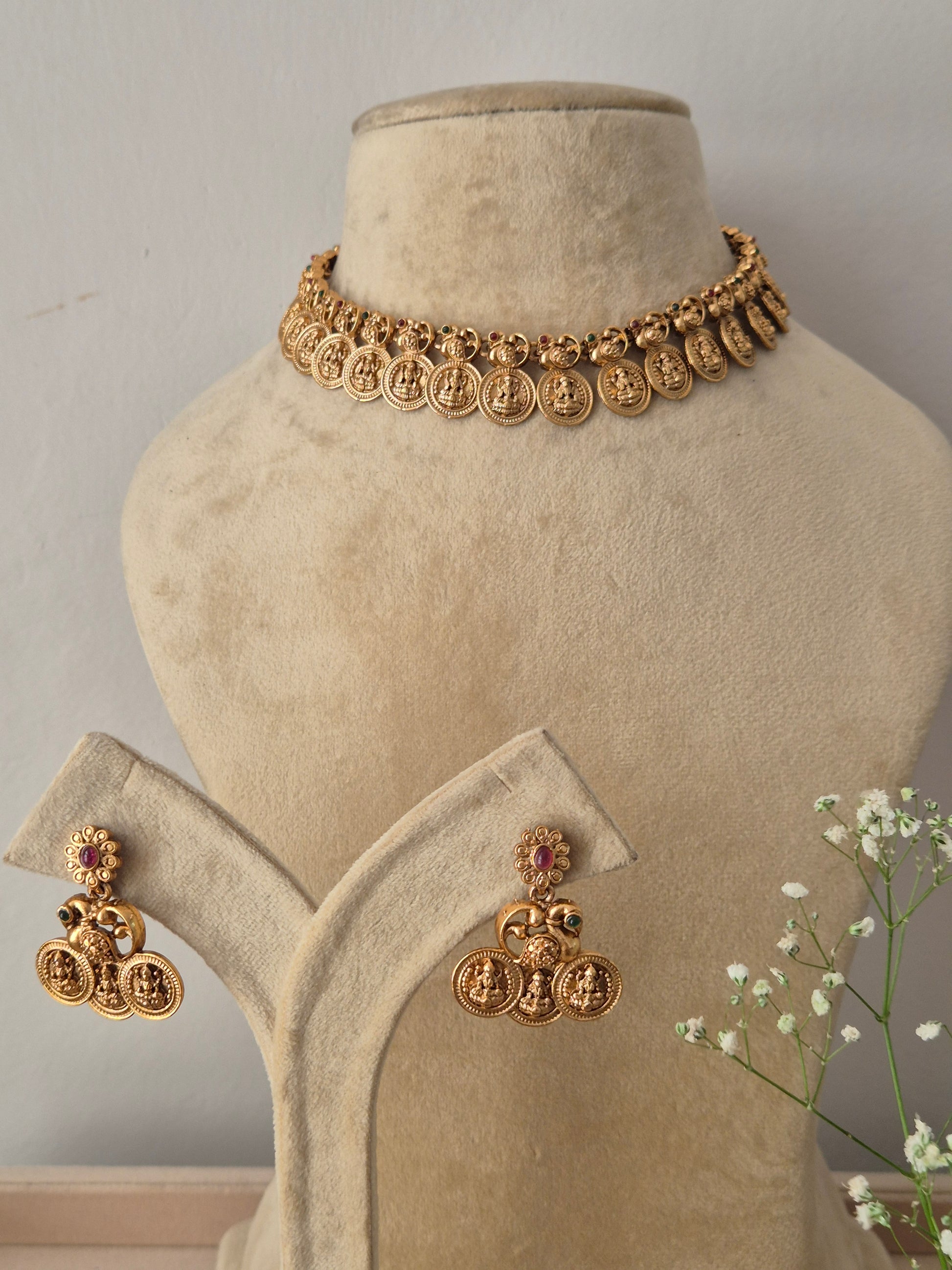 A gold-plated choker necklace featuring a repeating pattern of traditional coin-shaped motifs, with matching hanging jhumka earrings featuring pink accents by naitri.in