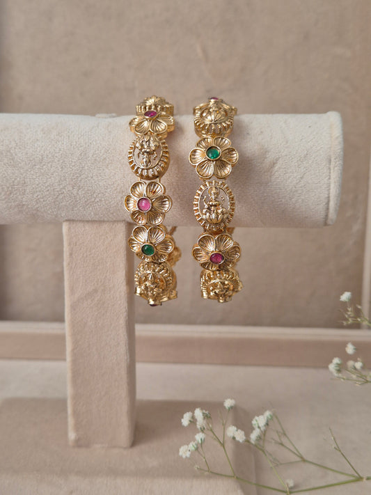 Set of 2 Maitri gold bangles by Naitri featuring intricate traditional floral engravings, accented with alternating ruby-red and emerald-green gemstones by naitri.in
