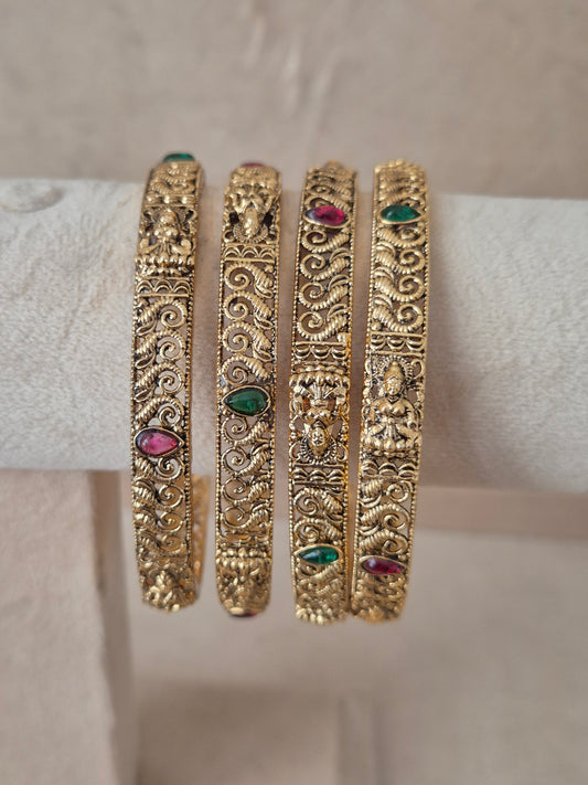Set of 4 gold bangles by Naitri featuring intricate ethnic floral patterns, embedded with alternating ruby-red and emerald-green gemstones by naitri.in