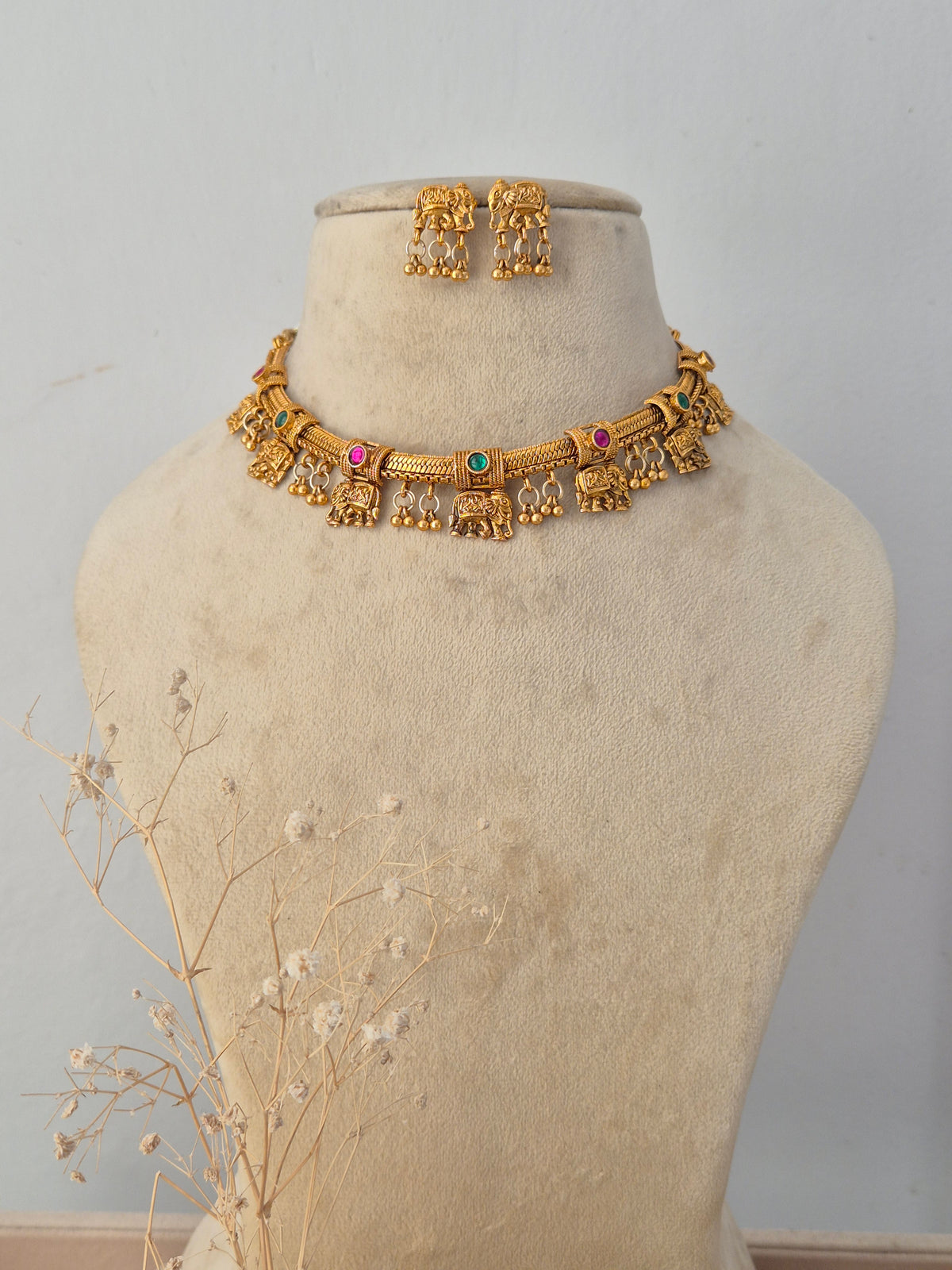 the handcrafted Mannu Necklace Set displayed on a neutral mannequin bust. The brass alloy choker is detailed with traditional ethnic motifs and vibrant gemstone accents. A pair of matching jhumka earrings rests neatly above the necklace, completing the traditional set by Naitri.in