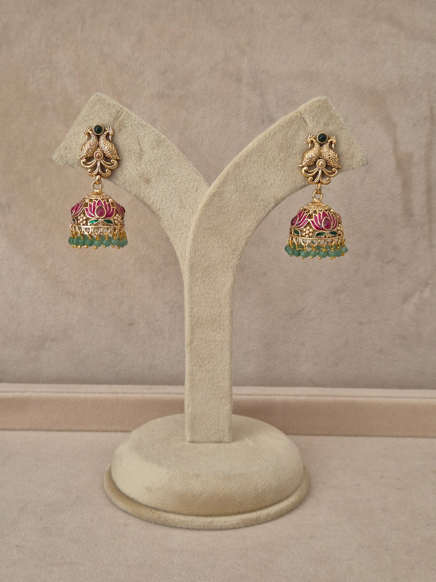 Pair of gold-plated Manorama Jhumka earrings hanging on a beige display stand, featuring detailed peacock motif studs with black stone accents and bell-shaped drops adorned with pink lotus flower enamel and green bead fringe by naitri.in