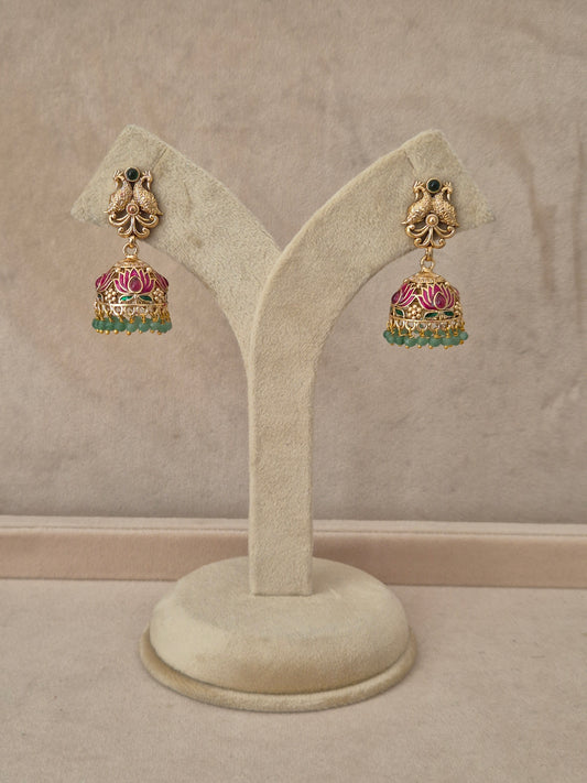 Pair of gold-plated Manorama Jhumka earrings hanging on a beige display stand, featuring detailed peacock motif studs with black stone accents and bell-shaped drops adorned with pink lotus flower enamel and green bead fringe by naitri.in
