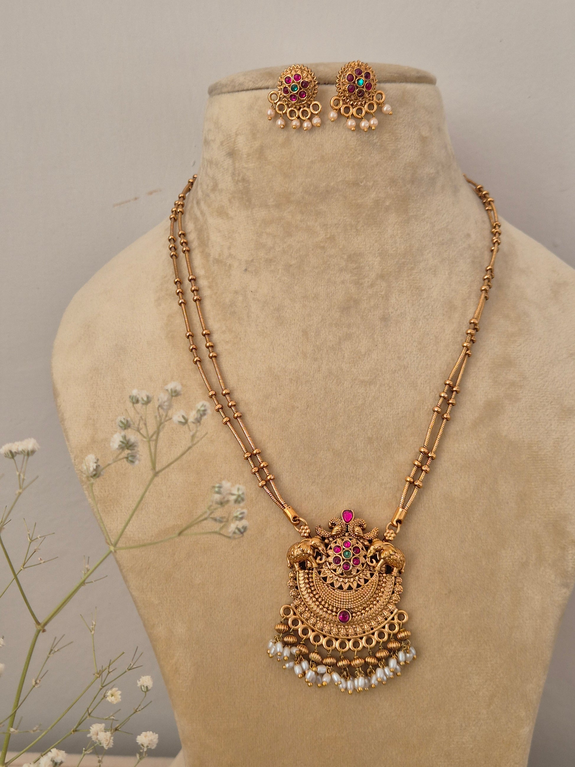 Handcrafted traditional gold-plated temple jewelry necklace featuring a delicate double-strand black and gold beaded chain, an intricate semicircular pendant with red and green stones, and dangling white pearl clusters with matching stud earring by naitri.in