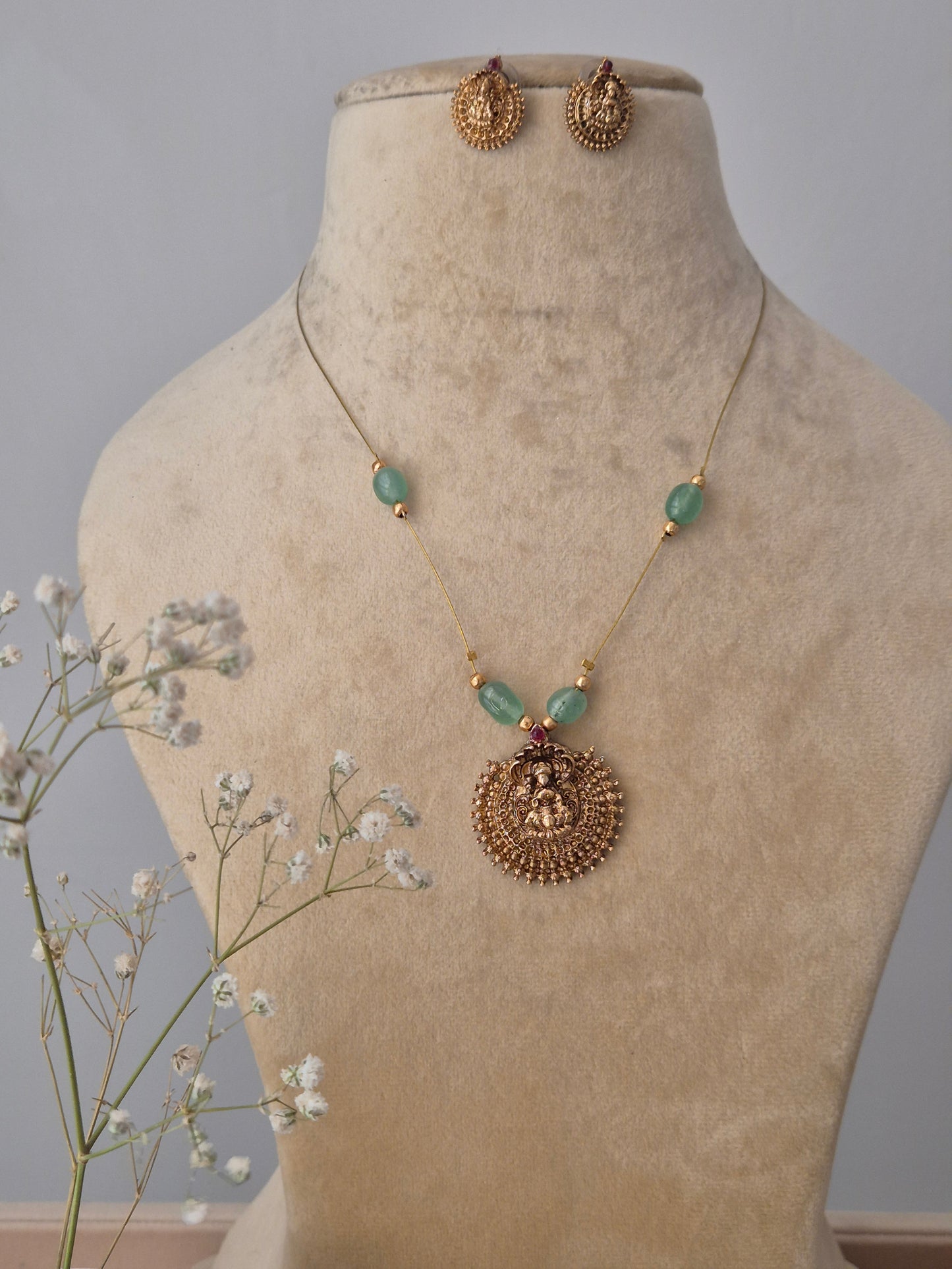 A gold-plated brass necklace with a circular deity-inspired pendant, featuring green stone accents and matching gold stud earrings  by naitri.in