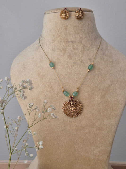 A gold-plated brass necklace with a circular deity-inspired pendant, featuring green stone accents and matching gold stud earrings  by naitri.in