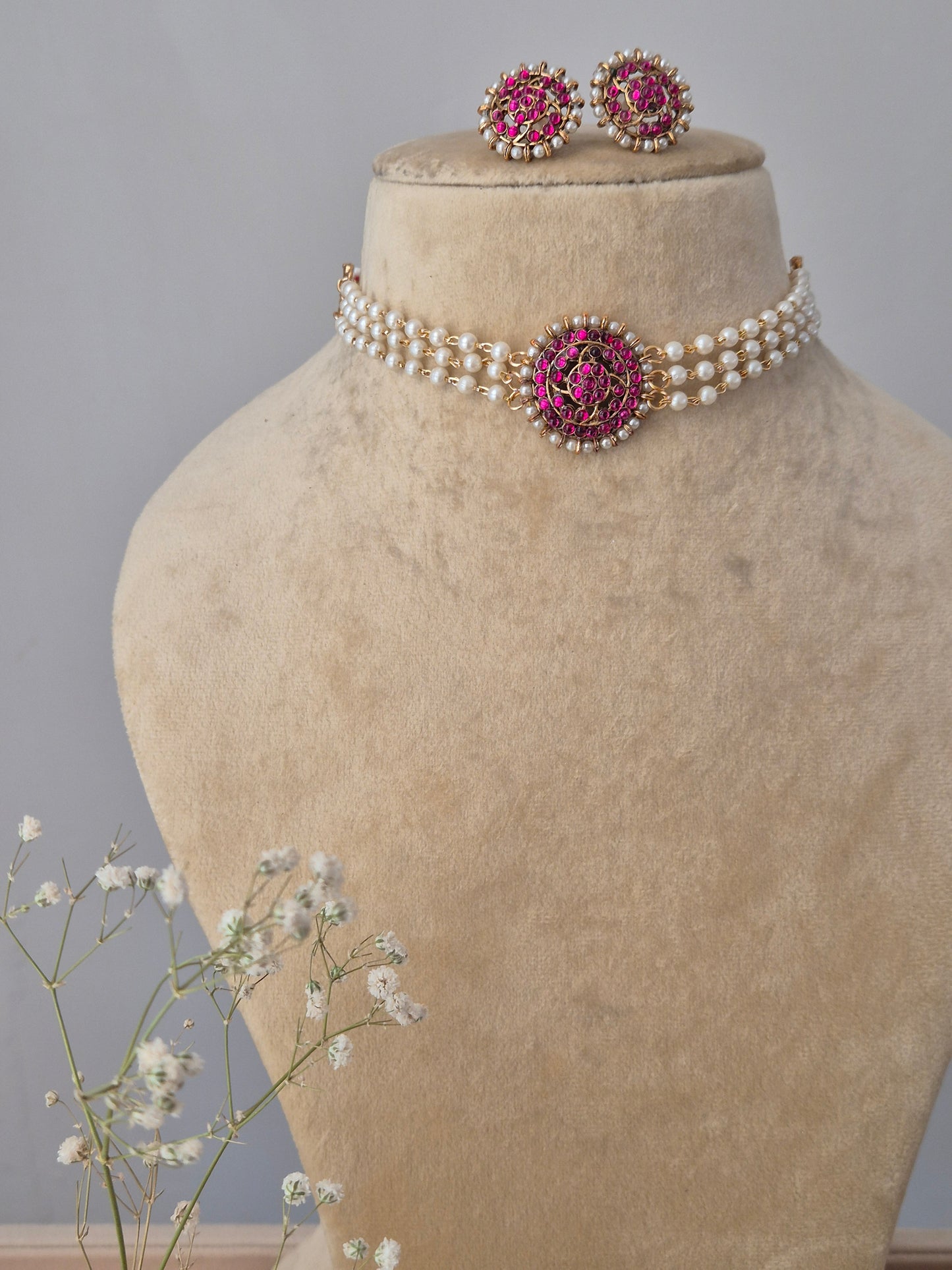 traditional Indian-style pearl choker with a ruby-pink stone floral medallion, displayed on a beige bust with delicate baby's breath flowers by naitri.in