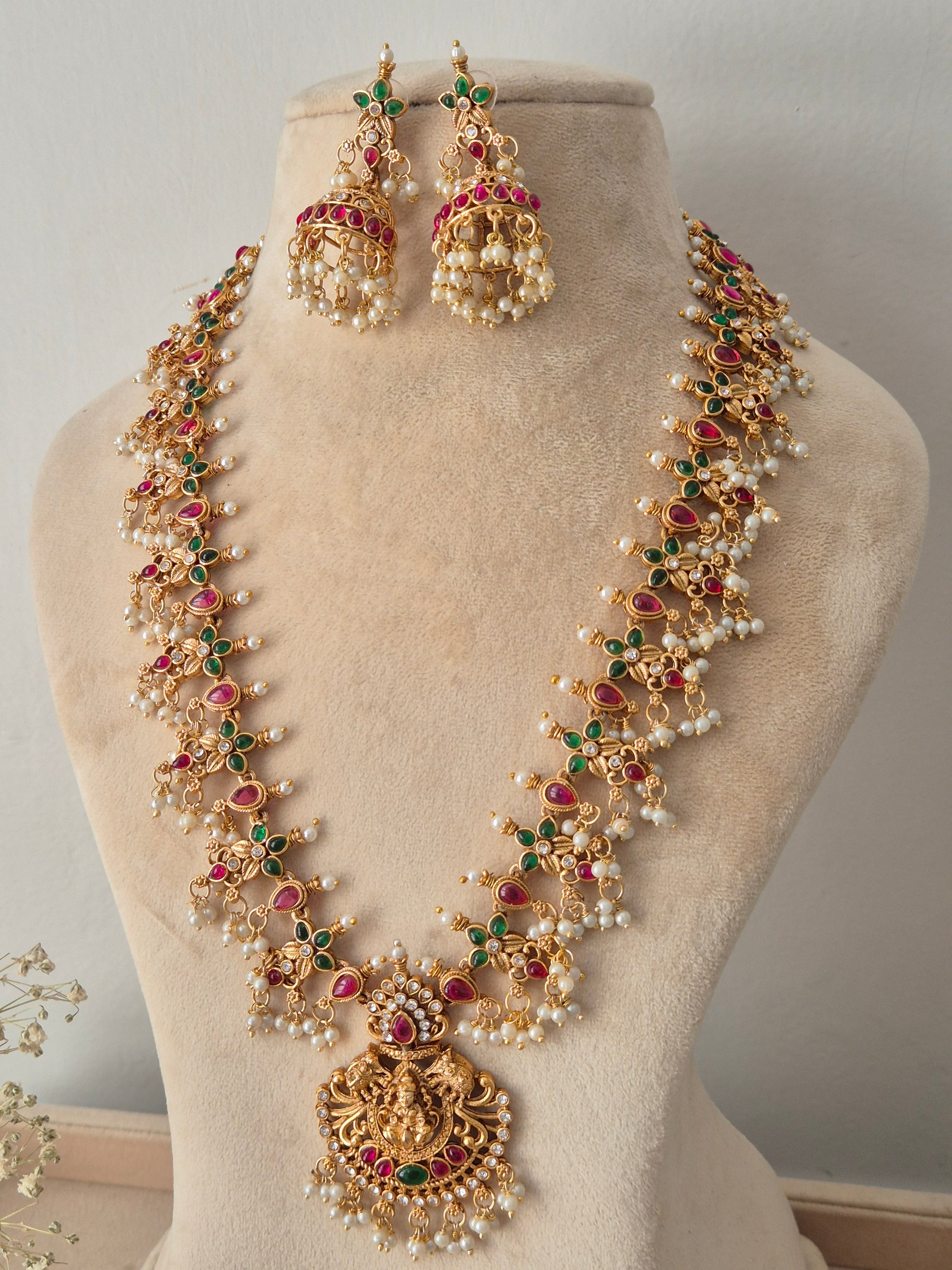 Traditional gold-tone temple necklace set featuring a large circular deity pendant, red-green stone work, hanging pearl clusters, and matching jhumka earrings By naitri.in