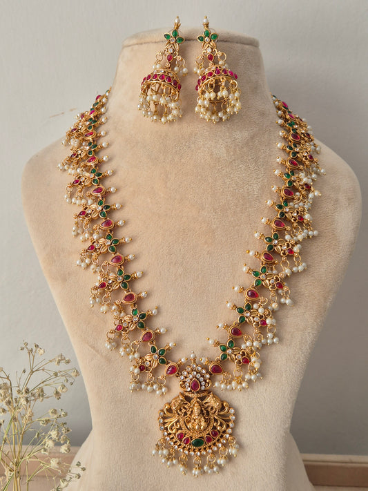 Gold-plated traditional Indian long necklace and earring set featuring red and green stone accents, pearl clusters, and an ornate deity pendant by naitri.in