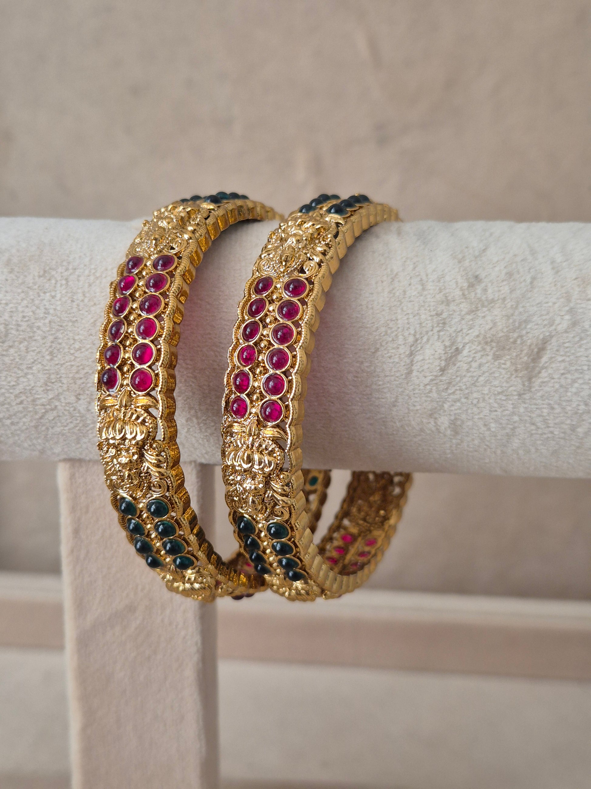 Pair of Naitri's Mitanshi gold bangles showcasing detailed ethnic craftsmanship, embedded with alternating vibrant pinkish-red and green accent stones, by naitri.in