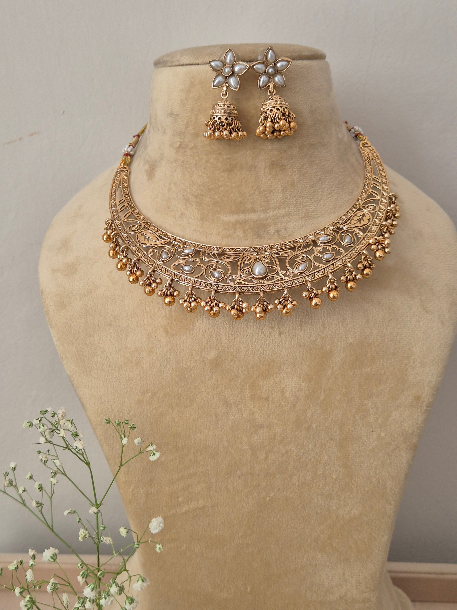 Handcrafted gold-plated temple jewelry choker necklace featuring intricate metal carvings, central white stone accents, and hanging gold pearl clusters, paired with matching white floral jhumka earrings by naitri.in
