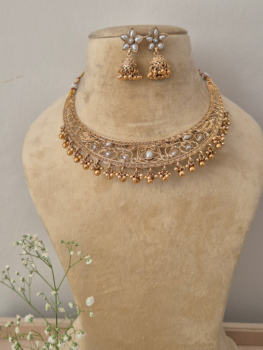Handcrafted gold-plated temple jewelry choker necklace featuring intricate metal carvings, central white stone accents, and hanging gold pearl clusters, paired with matching white floral jhumka earrings by naitri.in