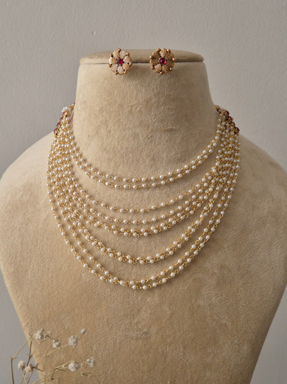 A ten-row graduated pearl strand necklace with pink gemstone accents at the clasps, accompanied by a pair of gold-toned floral-shaped stud earrings by naitri.in