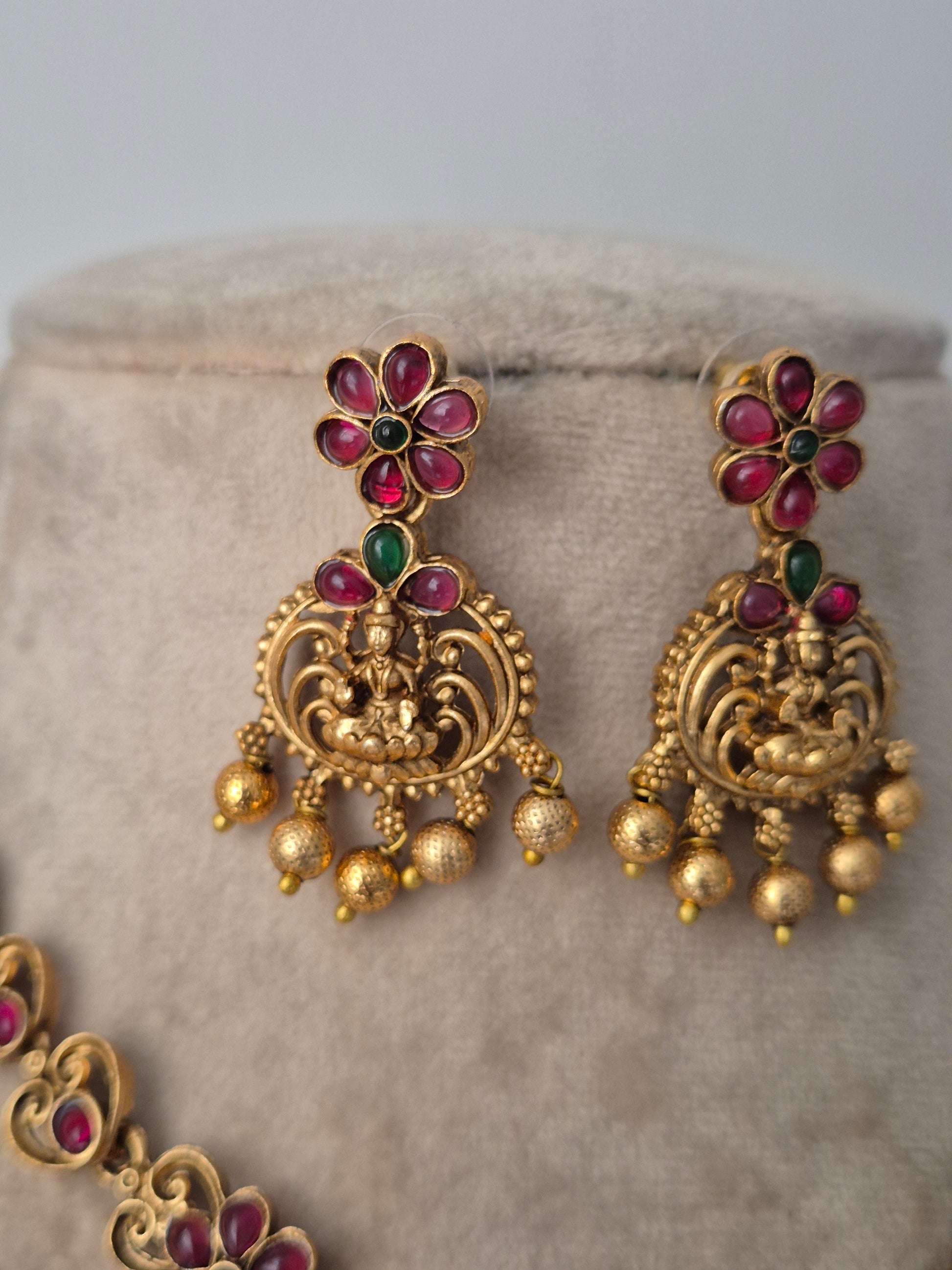 Gold earrings with red and green gemstones on a textured surface Temple earring by naitri.in