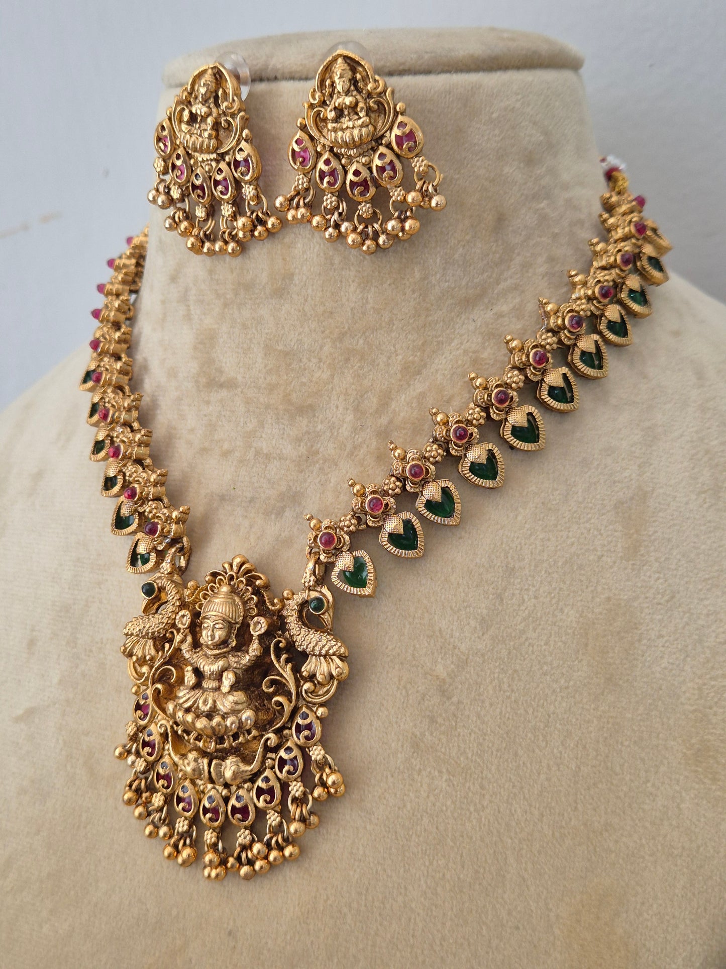 Modern Indian ethnic imitation jewelry set including a golden brass alloy necklace with intricate divine engravings, emerald green and ruby red stones, by naitri.in