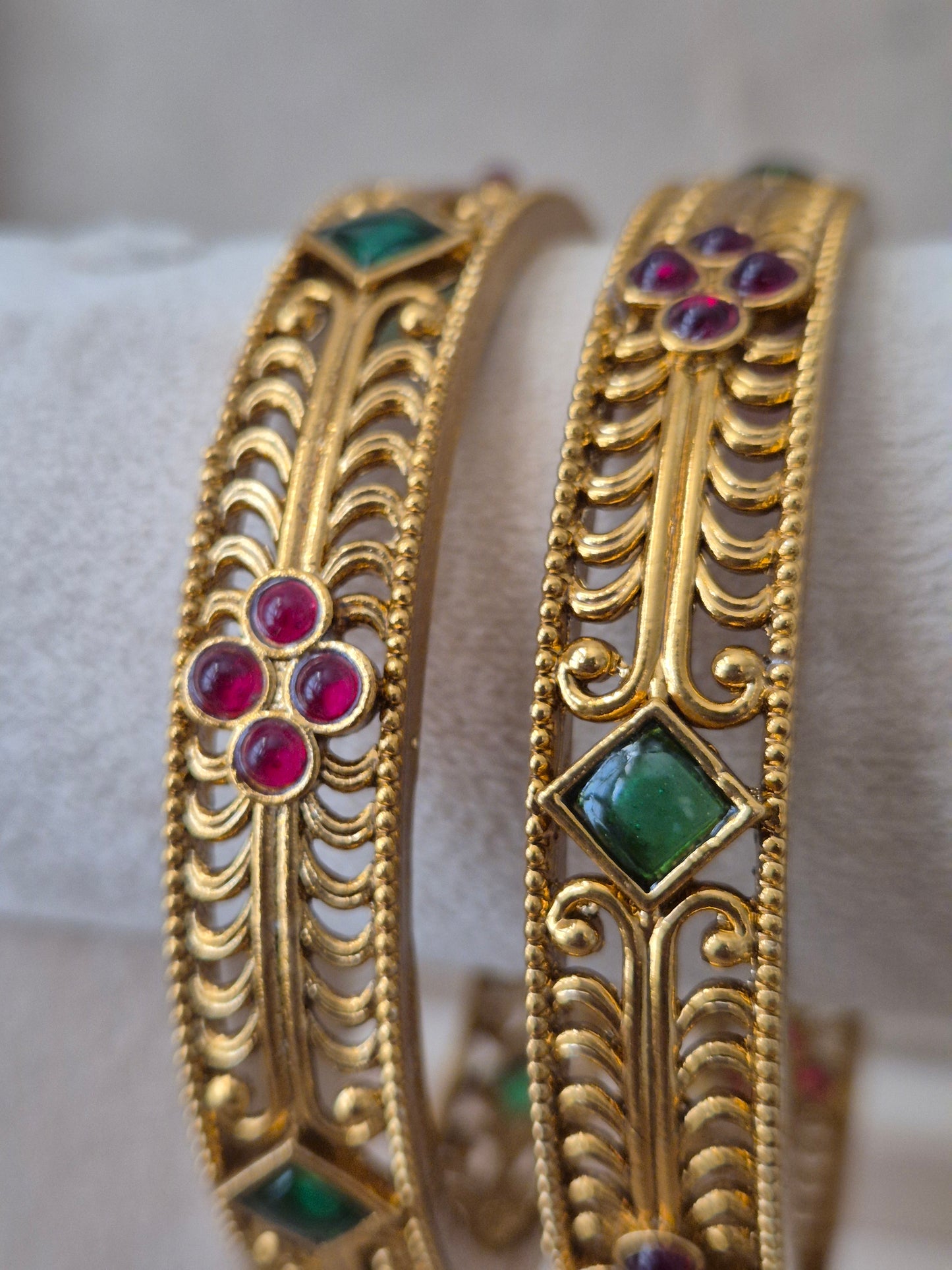 Two traditional Indian gold bangles from Naitri featuring detailed ethnic patterns, alternating ruby-red floral motifs, and emerald-green diamond-shaped gemstones by naitri.in