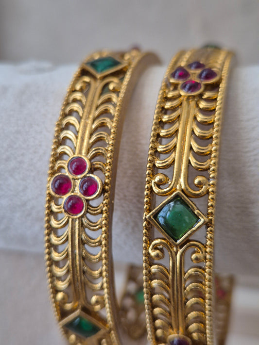 Two traditional Indian gold bangles from Naitri featuring detailed ethnic patterns, alternating ruby-red floral motifs, and emerald-green diamond-shaped gemstones by naitri.in