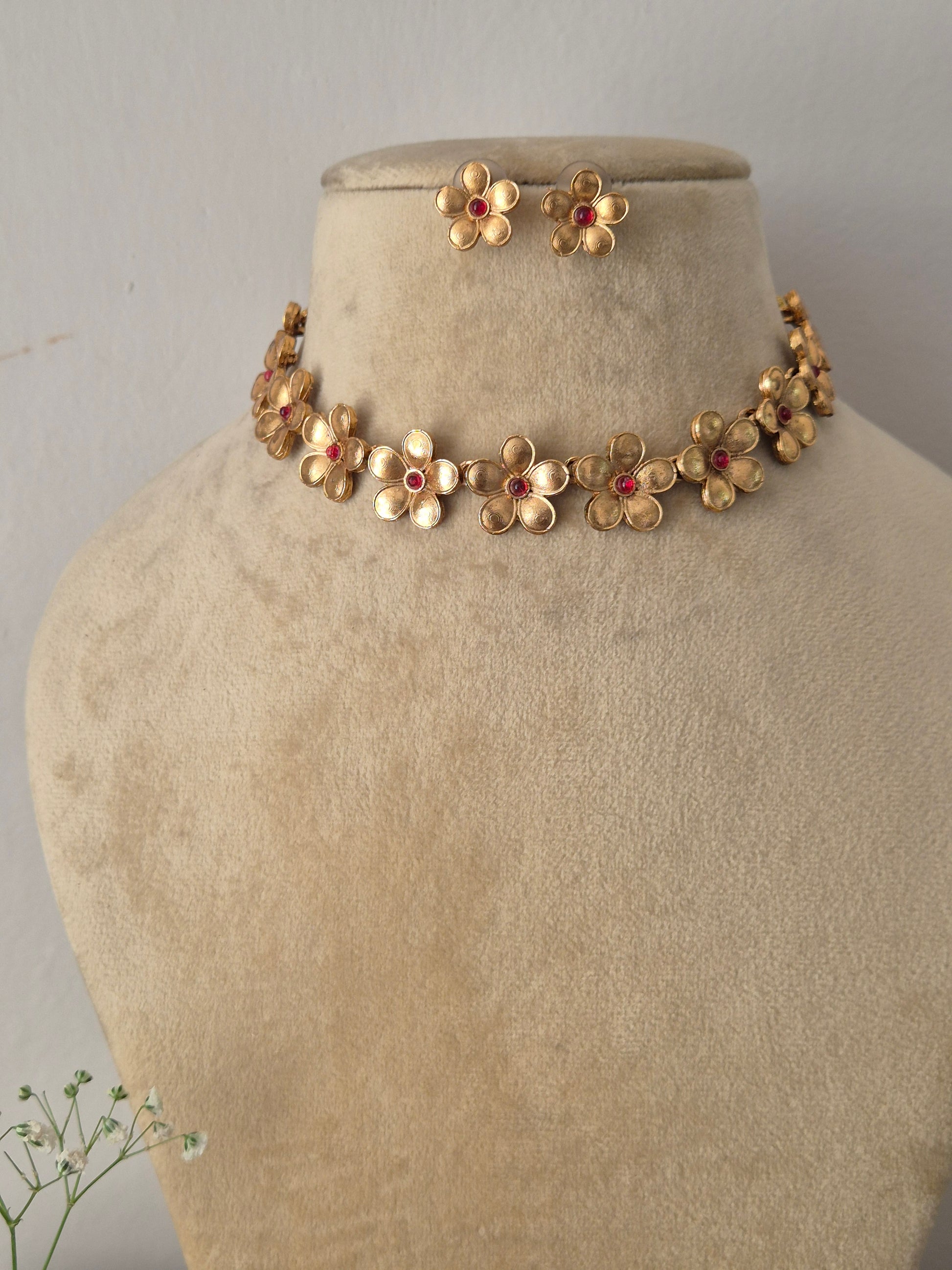 Modern Indian ethnic imitation jewelry set including a golden brass alloy choker necklace crafted with delicate floral motifs and pinkish-red stone centers, shown with matching flower earrings by naitri.in