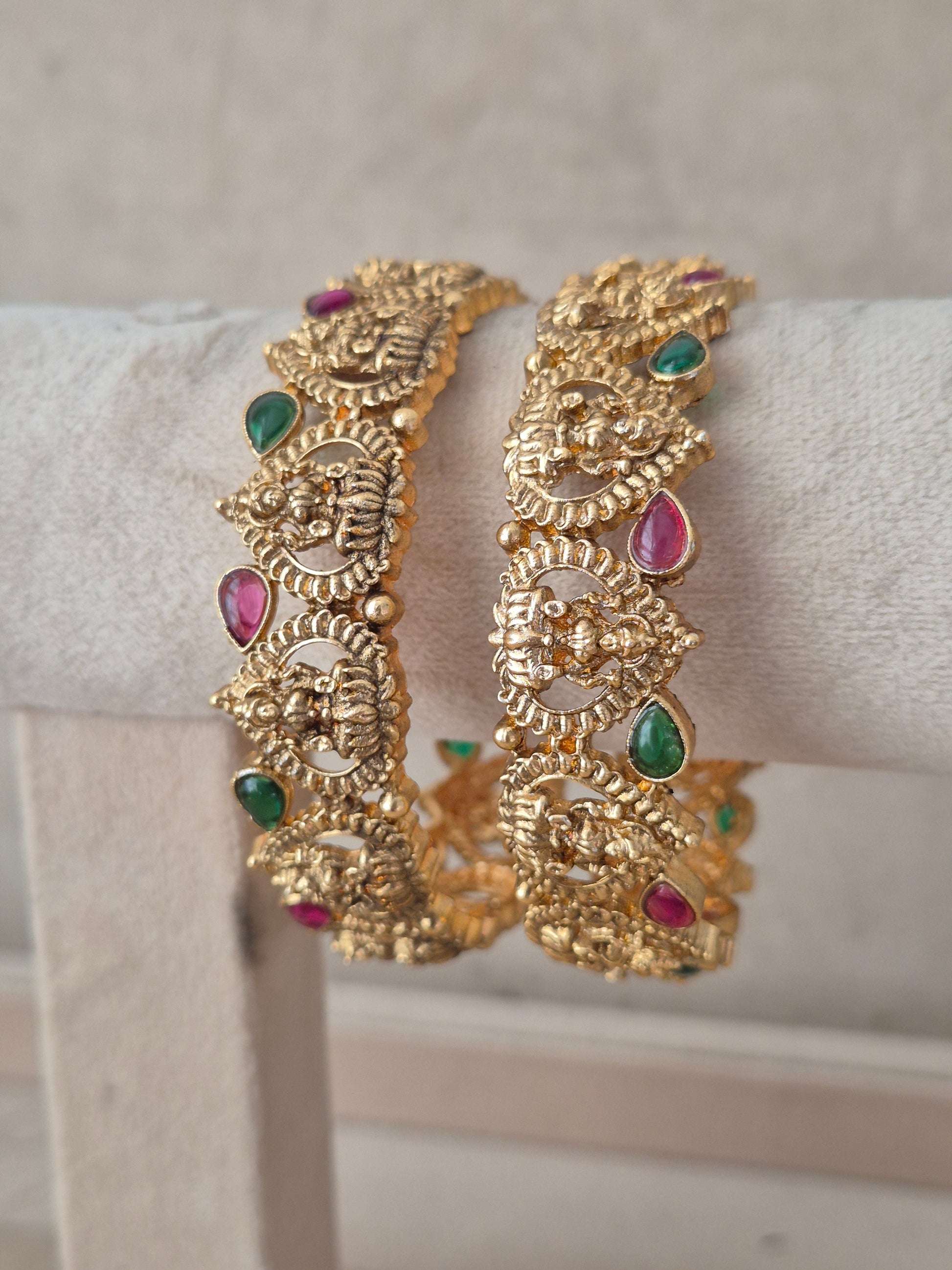 Pair of antique gold-toned traditional Indian bangles featuring intricate deity motifs, studded with green and ruby-red drop stones  by naitri.in
