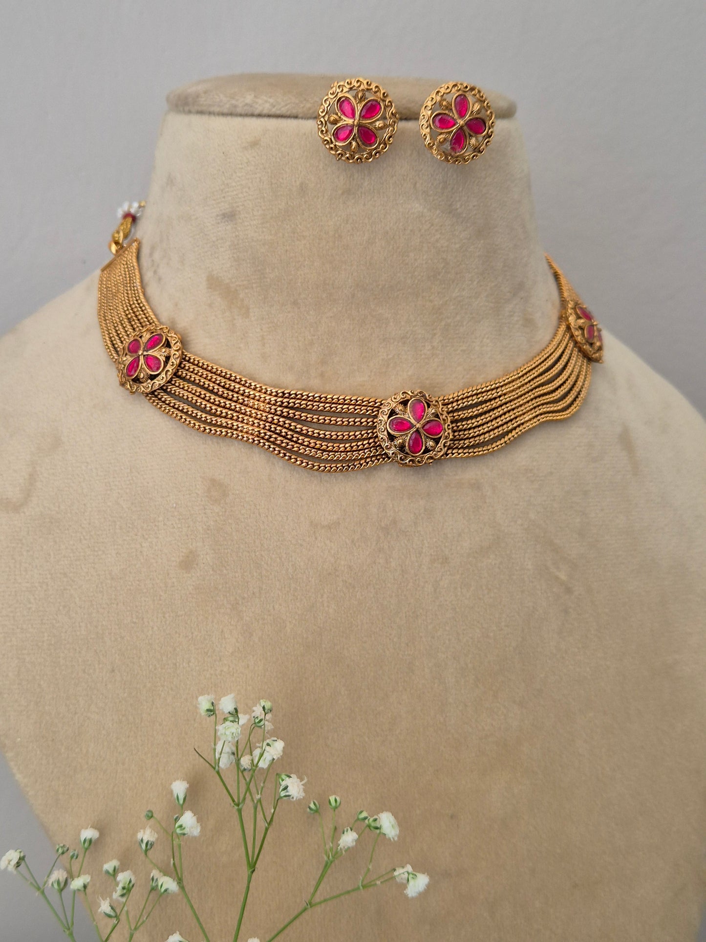 Modern Indian imitation jewelry set including a multi-layered golden strand choker necklace with a central pink stone floral motif and two matching round floral earrings by naitri.in