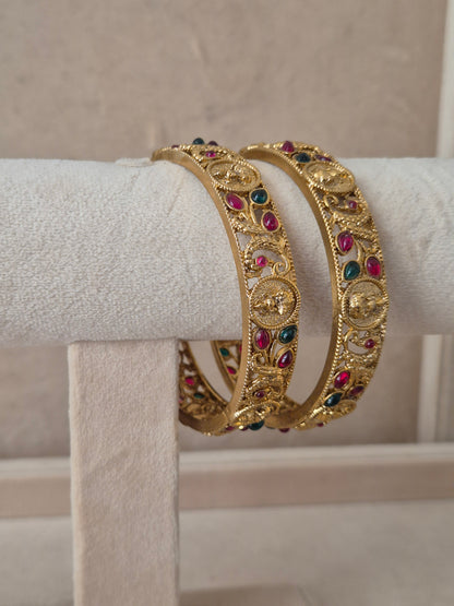 Two traditional gold-plated ethnic bangles with detailed textured patterns and inlaid red and green gemstones by naitri.in