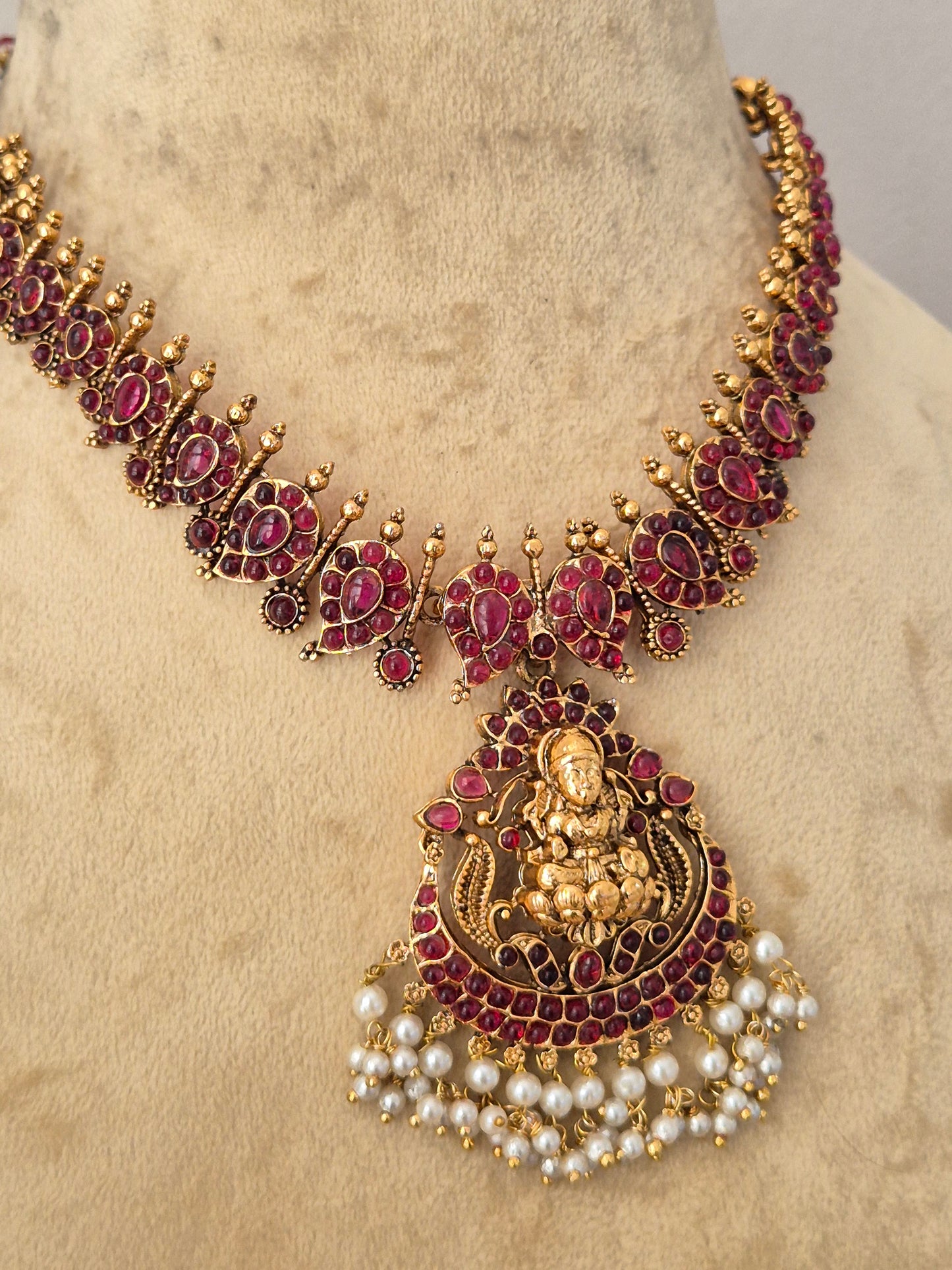 An ornate traditional Indian jewelry set featuring a gold-plated necklace and matching pendant decorated with deep red stones and white faux pearls, by naitri.in