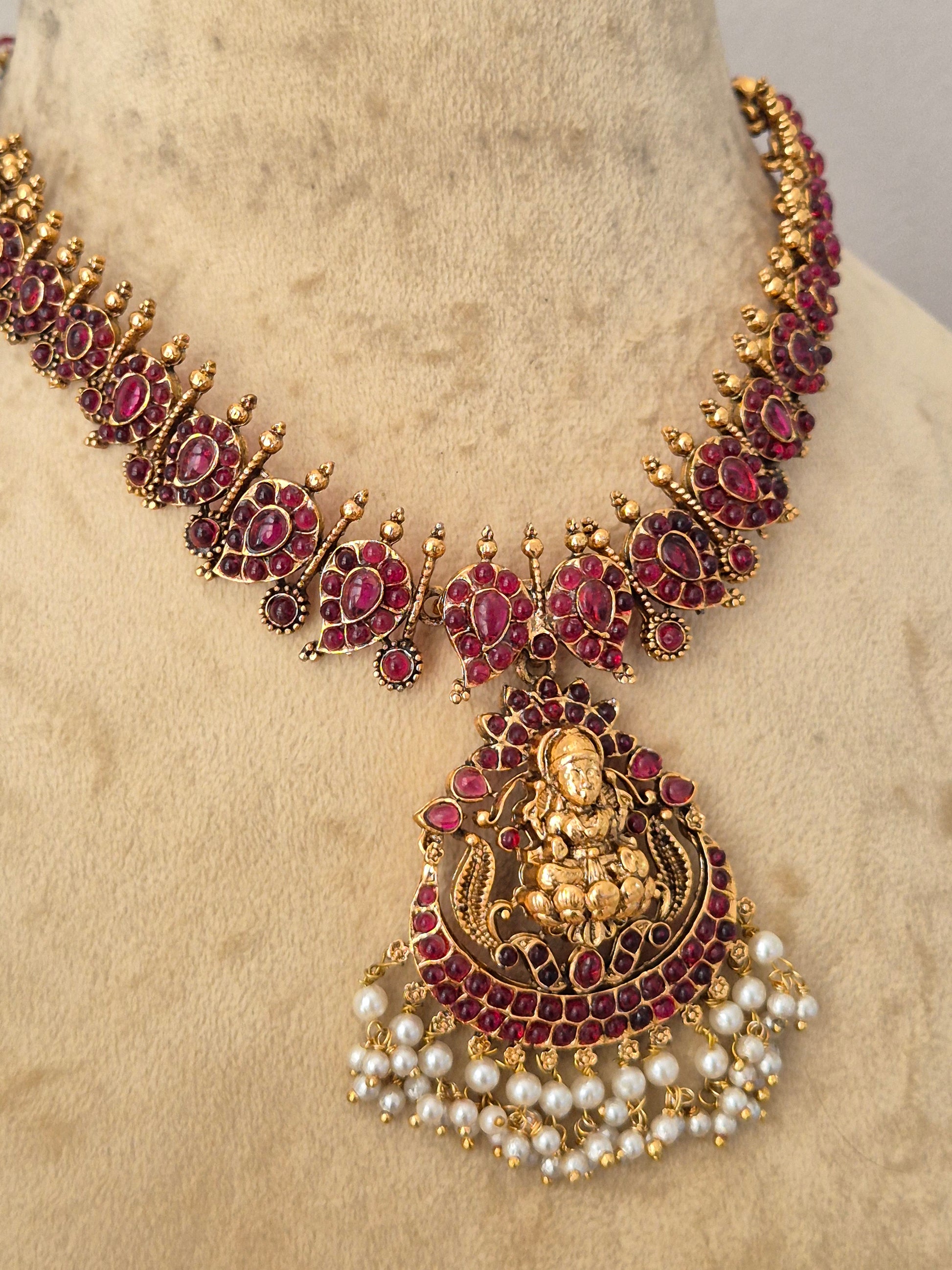 An ornate traditional Indian jewelry set featuring a gold-plated necklace and matching pendant decorated with deep red stones and white faux pearls, by naitri.in