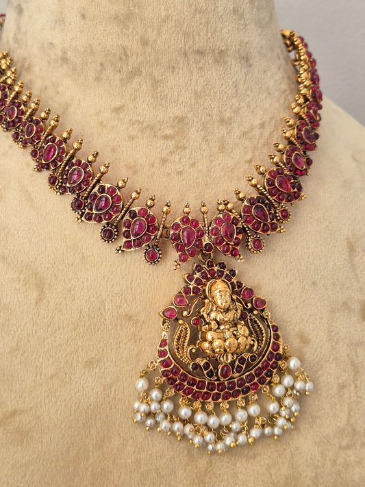 An ornate traditional Indian jewelry set featuring a gold-plated necklace and matching pendant decorated with deep red stones and white faux pearls, by naitri.in