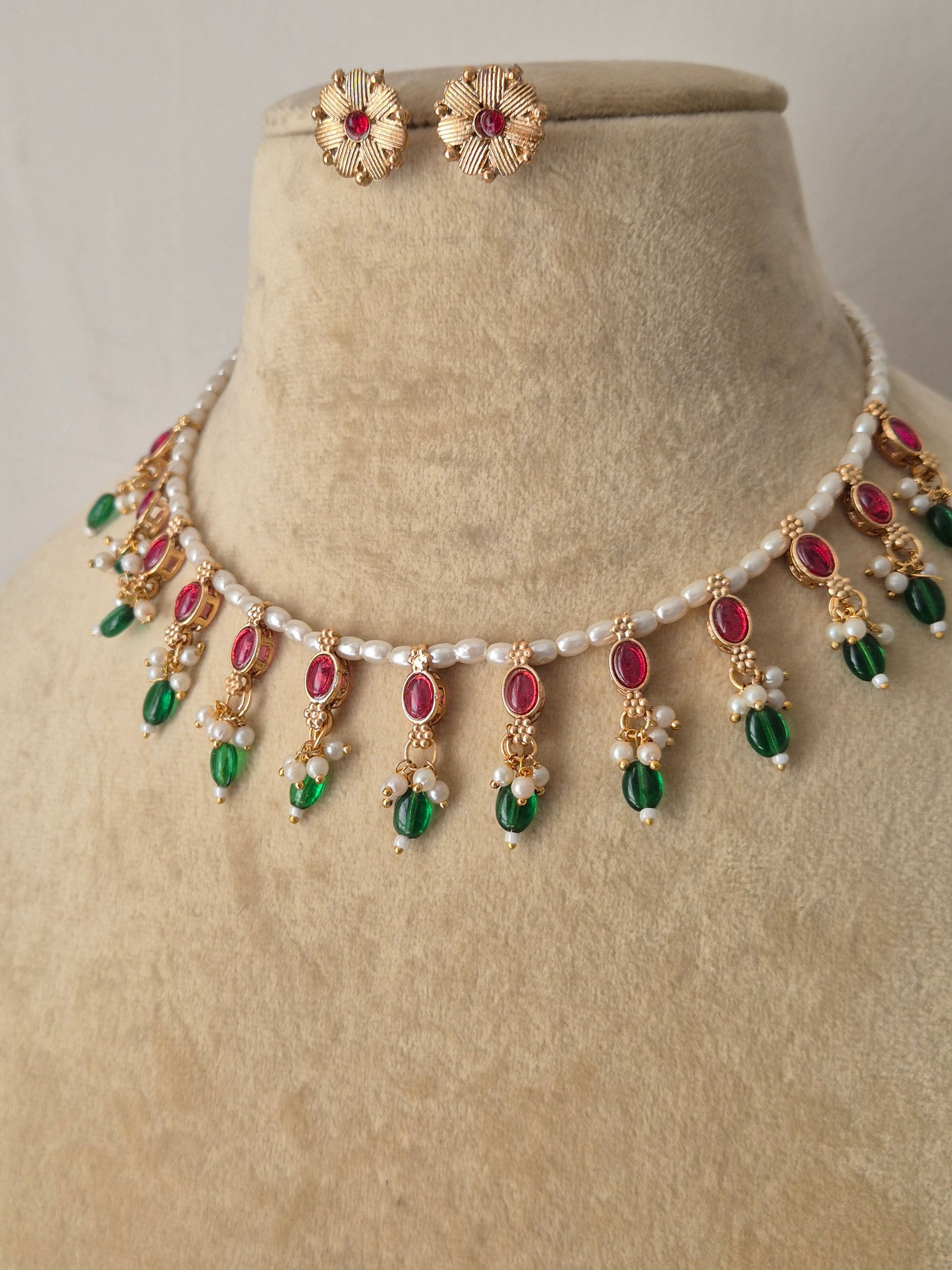 Traditional Indian antique-finish brass jewelry set showcasing a dainty pearl collar layout, vibrant pinkish-red and green gemstone charms, and coordinating floral-stamped stud earrings from the Naitri collection by naitri.in