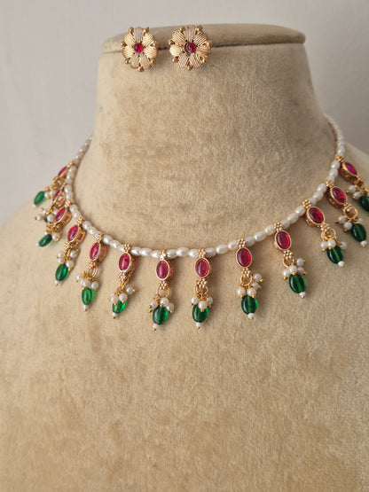 Traditional Indian antique-finish brass jewelry set showcasing a dainty pearl collar layout, vibrant pinkish-red and green gemstone charms, and coordinating floral-stamped stud earrings from the Naitri collection by naitri.in
