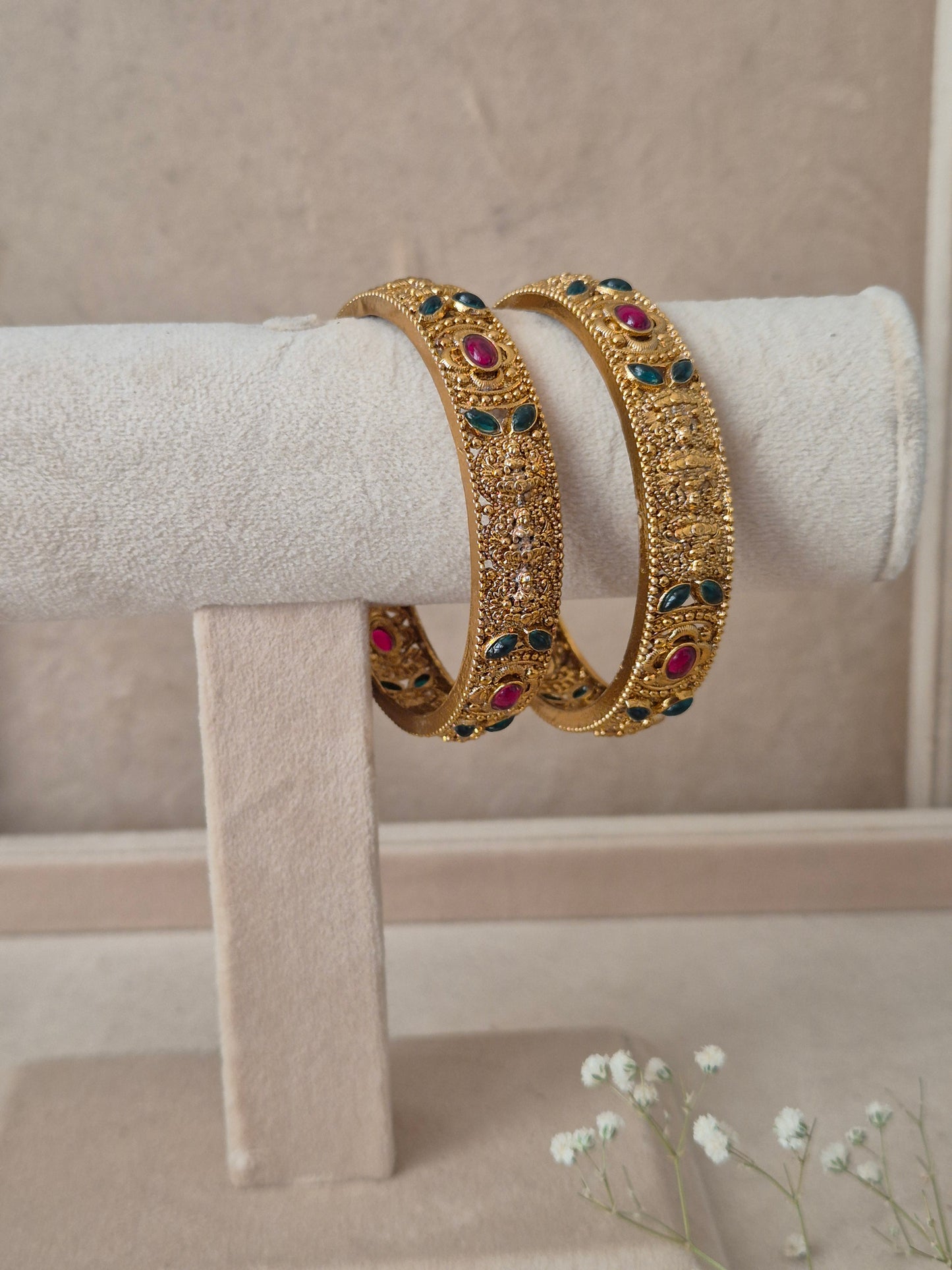 Two traditional gold-plated ethnic bangles with detailed textured patterns and inlaid red and green gemstones by naitri.in