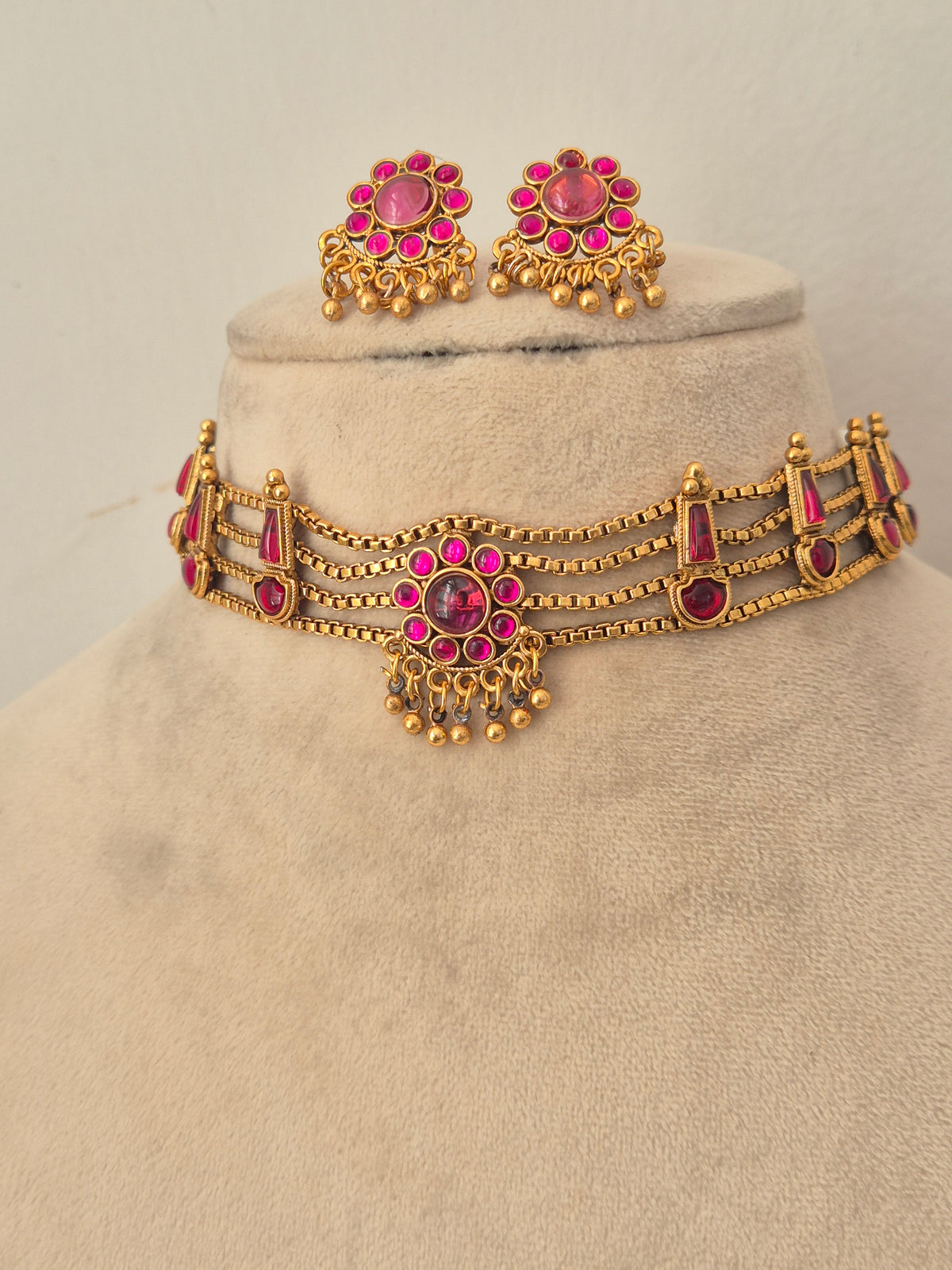 Antique gold-toned choker necklace showcasing an intricate metallic floral band, vibrant pink stone clusters, and delicate hanging gold bead drops, shown with coordinating round stud earrings by naitri.in