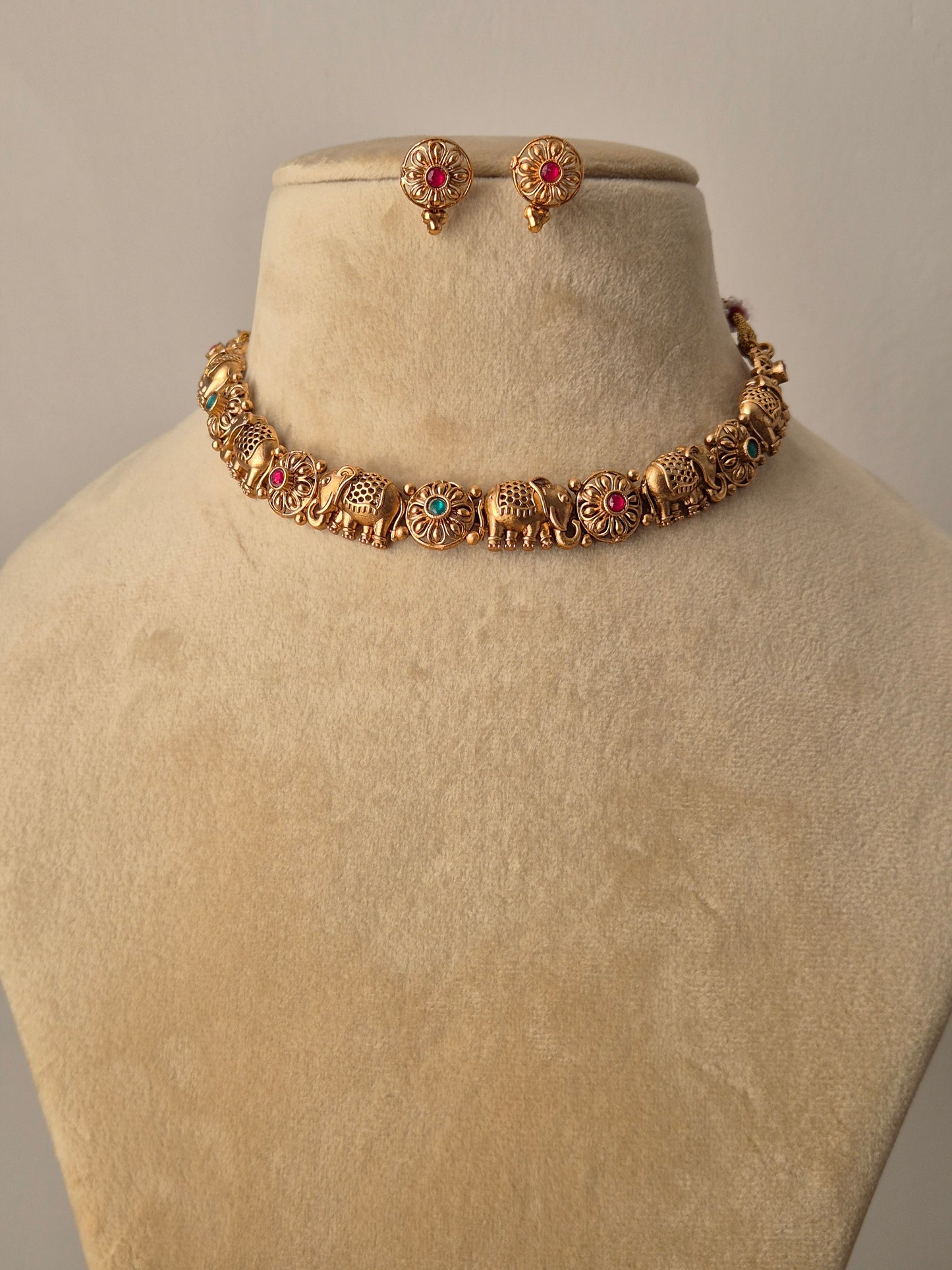 Close-up of the Disha Necklace Set: a golden brass choker featuring a row of detailed elephant figures with small red and green stones, paired with matching floral-style studs by naitri.in