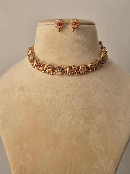 Close-up of the Disha Necklace Set: a golden brass choker featuring a row of detailed elephant figures with small red and green stones, paired with matching floral-style studs by naitri.in