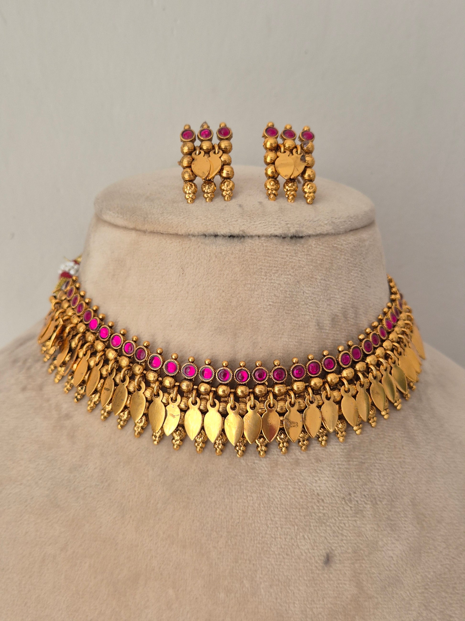 Gold-toned traditional jewelry set including a choker necklace with dangling leaf-shaped charms, matching earrings, and small pink stones throughout by naitri.in