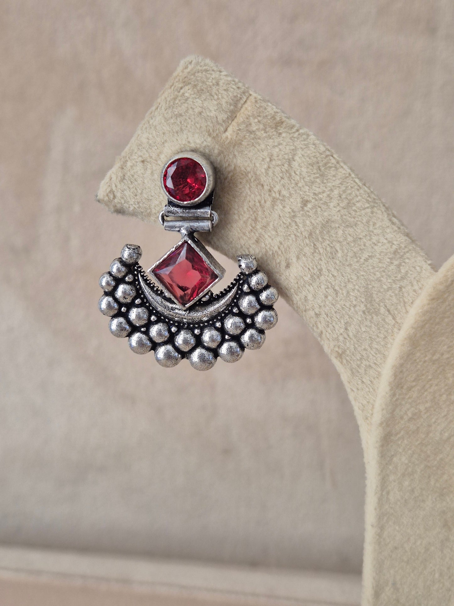 Silver earring with red gemstones on a beige stone surface Naitri premium Falguni oxidized silver earrings featuring red stones from the Silver Jewellery collection by naitri.in