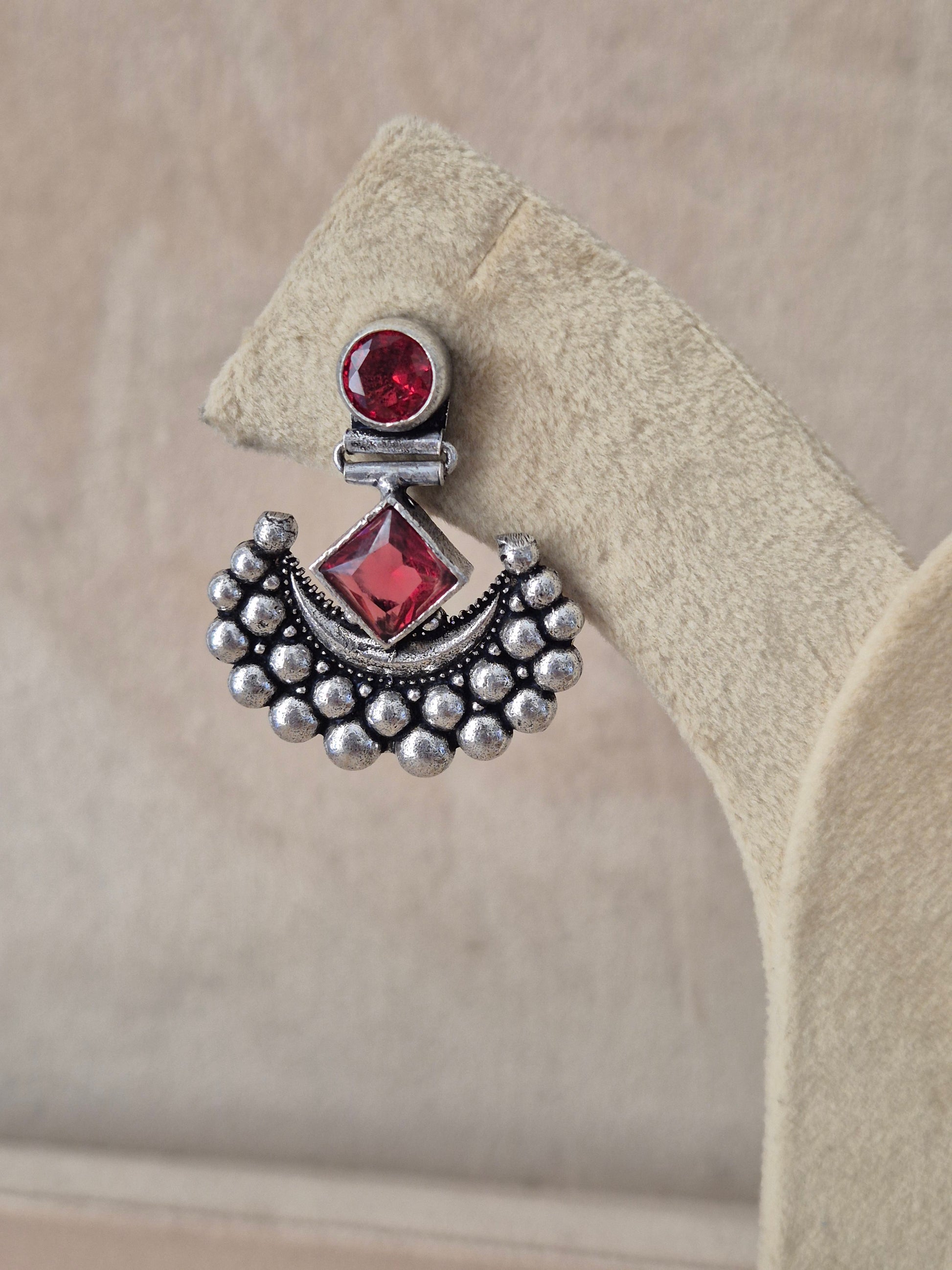 Silver earring with red gemstones on a beige stone surface Naitri premium Falguni oxidized silver earrings featuring red stones from the Silver Jewellery collection by naitri.in
