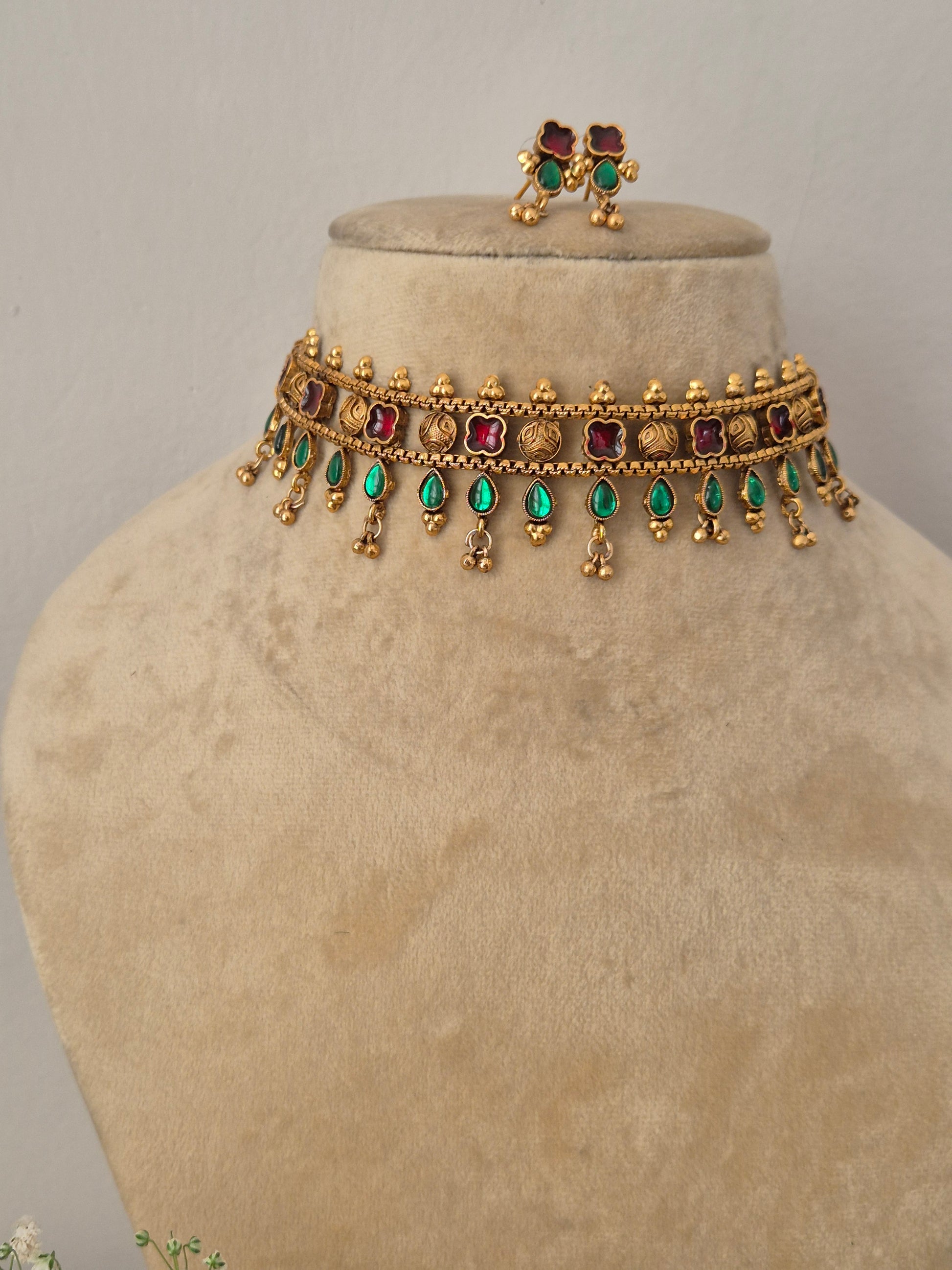 Modern Indian ethnic imitation jewelry set including a golden brass alloy choker necklace embedded with ruby red and emerald green stones, shown with matching small stud earrings by naitri.in