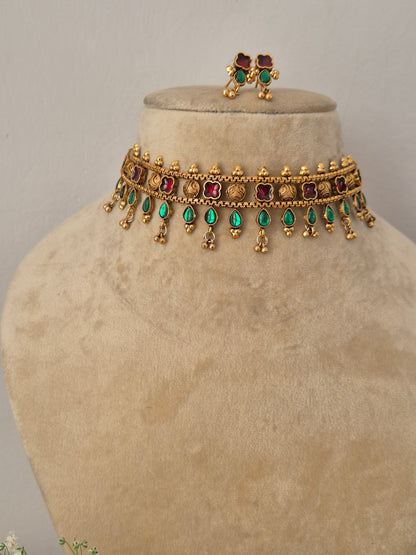 Modern Indian ethnic imitation jewelry set including a golden brass alloy choker necklace embedded with ruby red and emerald green stones, shown with matching small stud earrings by naitri.in