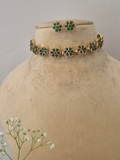 Modern Indian imitation jewelry set including a golden brass alloy choker necklace embedded with vibrant green stones in a repeating floral pattern, shown with matching studs by naitri.in