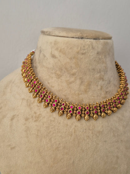 Gold-toned traditional Indian choker necklace with pink gemstone accents,  The choker features a row of vibrant pink gemstones bordered by intricate, textured gold-plated leaf-shaped drops by naitri.in