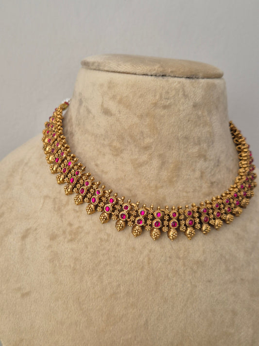 Gold-toned traditional Indian choker necklace with pink gemstone accents,  The choker features a row of vibrant pink gemstones bordered by intricate, textured gold-plated leaf-shaped drops by naitri.in