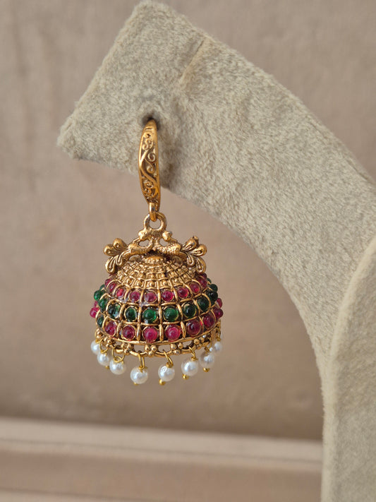 Close-up of Naitri Hemakshi premium gold ethnic earrings showcasing traditional Indian temple jewelry craftsmanship with vibrant ruby red and emerald green stone accents and delicate pearl bead fringe by naitri.in