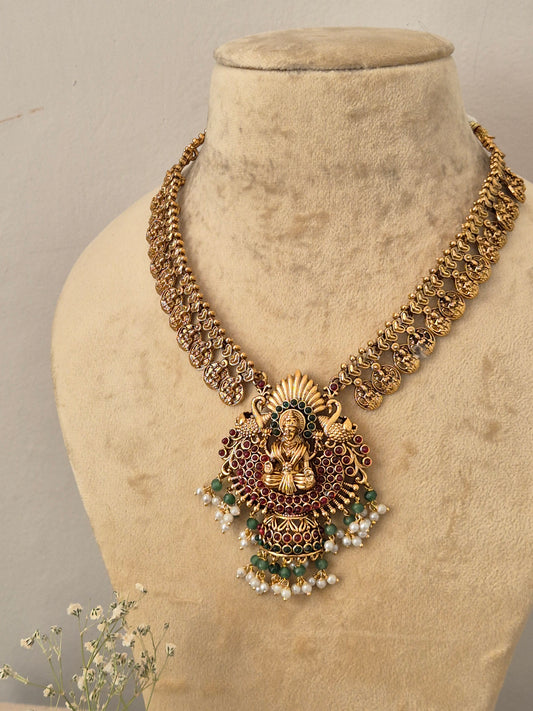 An ornate gold-toned traditional necklace the necklace features a detailed central pendant with a deity motif, red and green enamel accents, and small white pearl-like beads, by naitri.in
