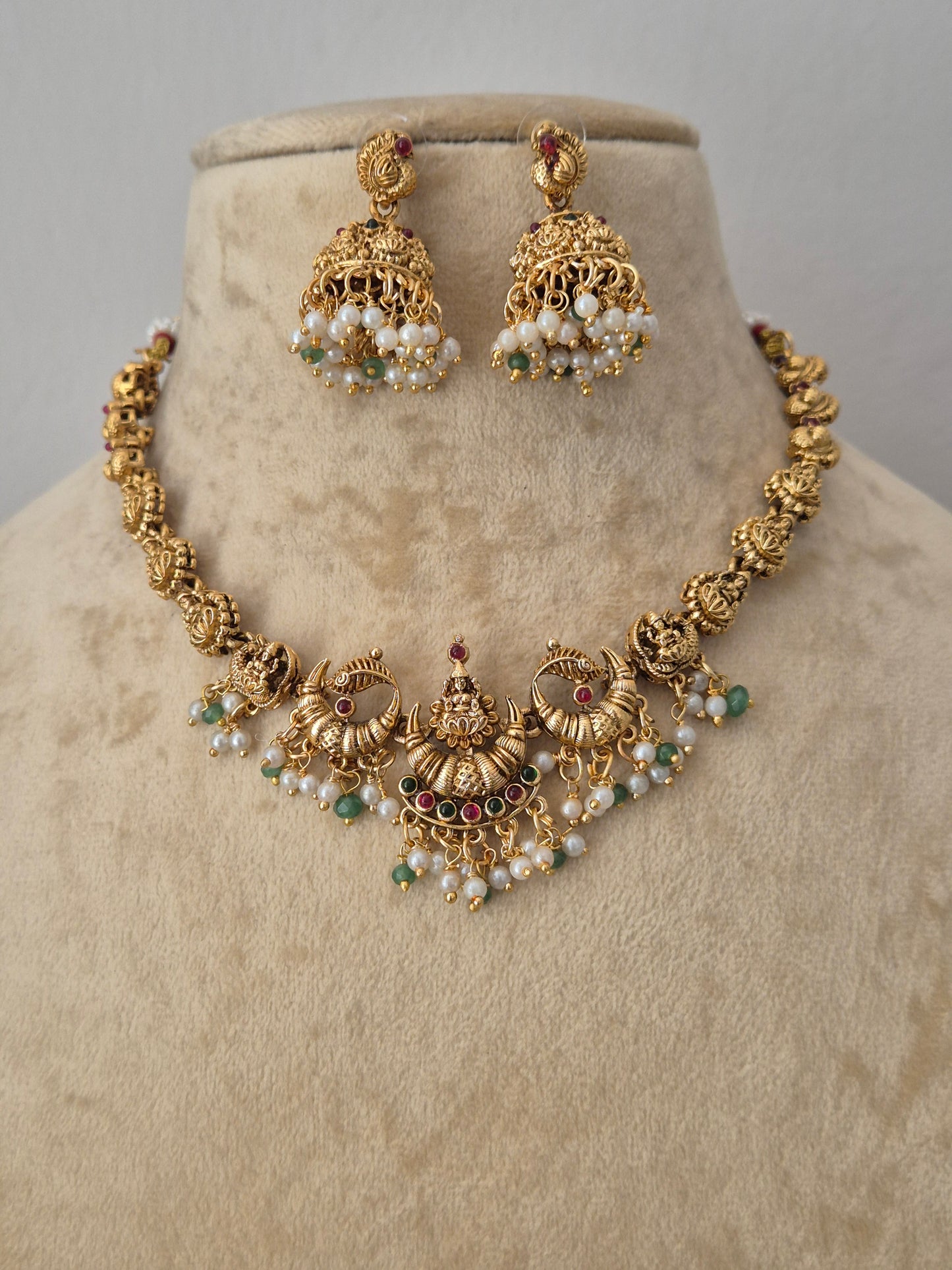 Gold-toned traditional Indian jewelry set featuring a jhumka earrings pair the pieces are decorated with white pearl-like beads, small green stones, and tiny red stones by naitri.in