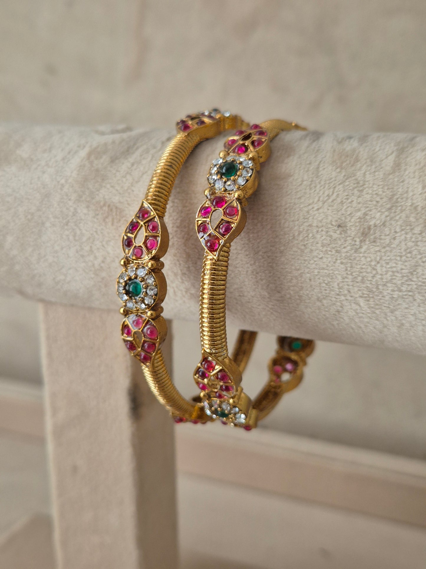A pair of gold-plated bangle bracelets with intricate floral engravings set with pink, green, and white stones, by naitri.in