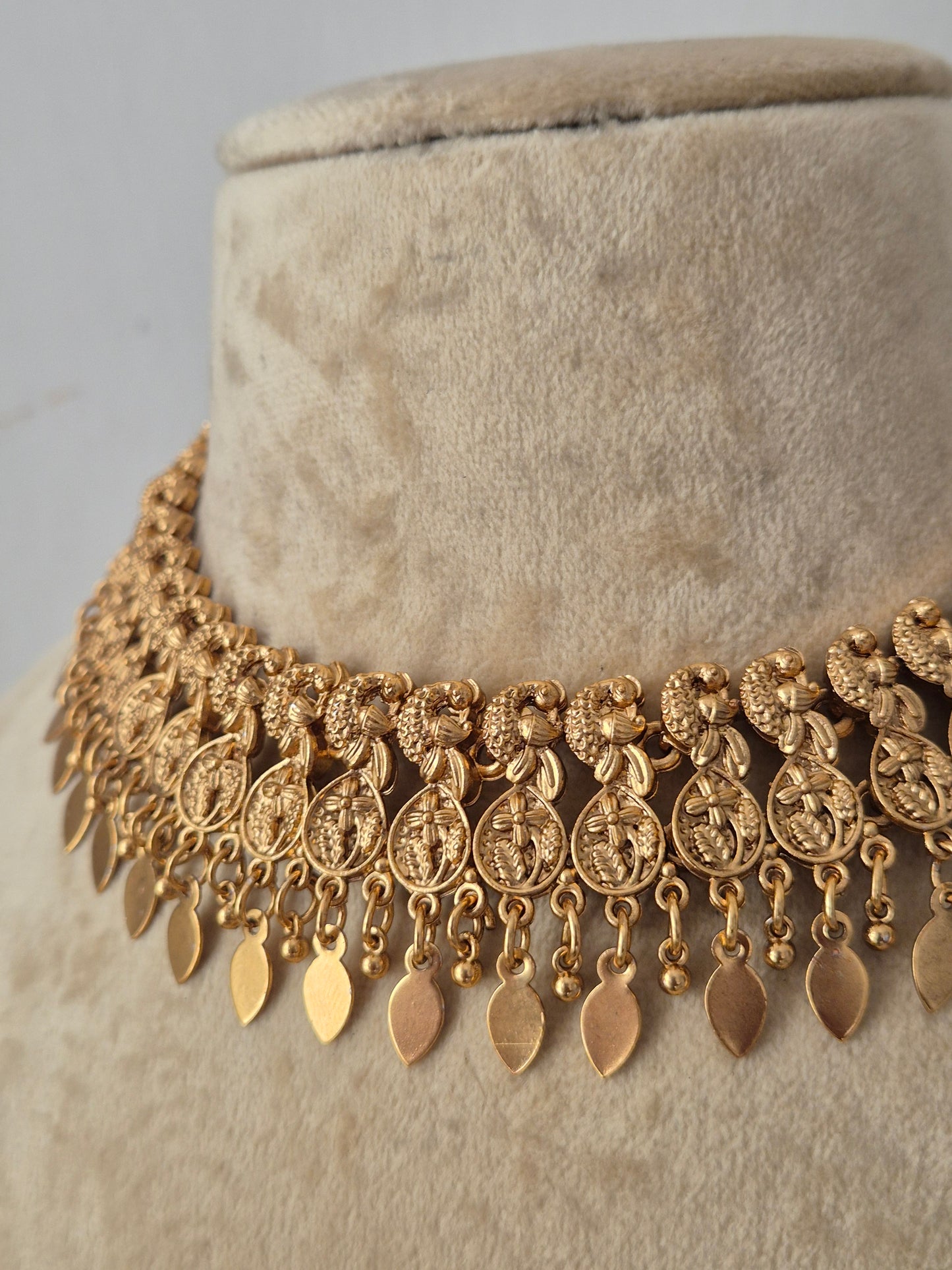 An ornate gold-toned statement necklace features a wide collar of textured floral and medallion motifs with dangling leaf-shaped charms by naitri.in