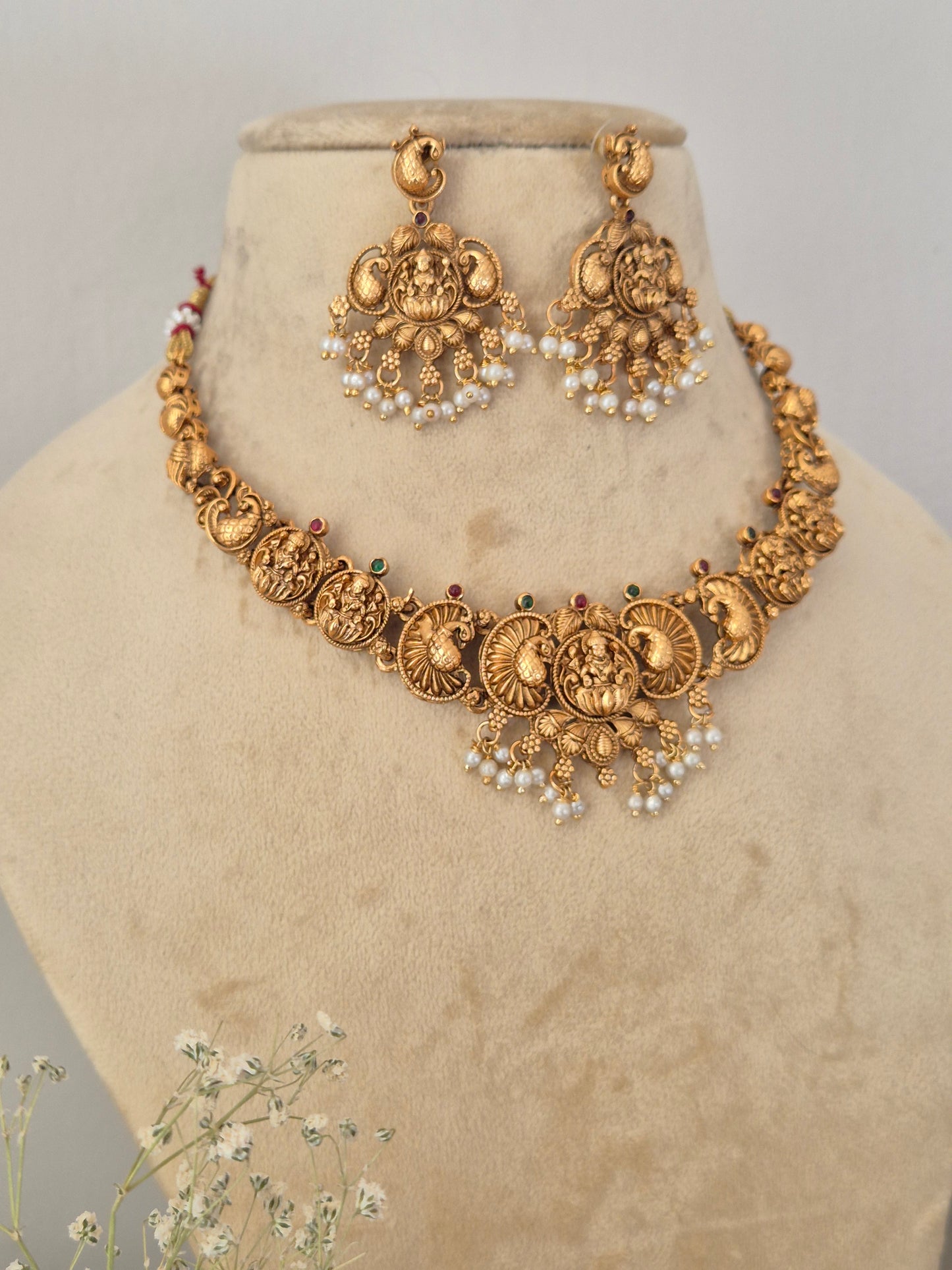 An ornate gold-toned traditional necklace and matching earrings Traditional gold-toned temple jewelry choker set with intricate circular motifs, goddess engravings, and white faux pearl drops, shown with matching dangle earrings by naitri.in