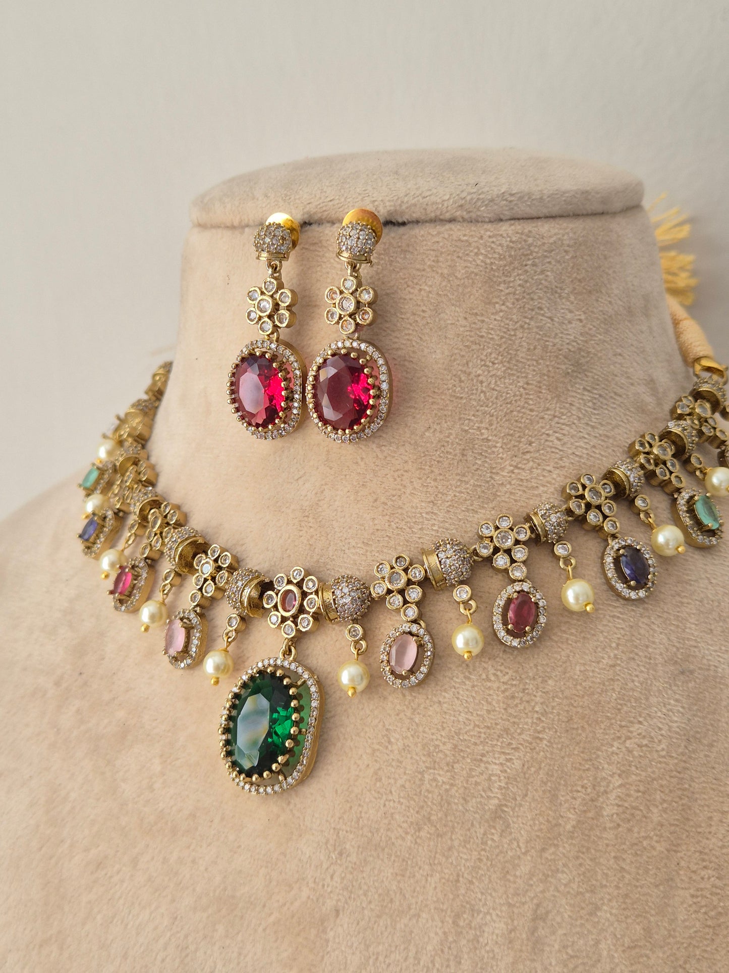 An ornate gold-toned traditional necklace set with matching earrings, the necklace features multiple oval and round gemstone settings in pink, green, purple, and blue, accented with small white faux pearls by naitri.in